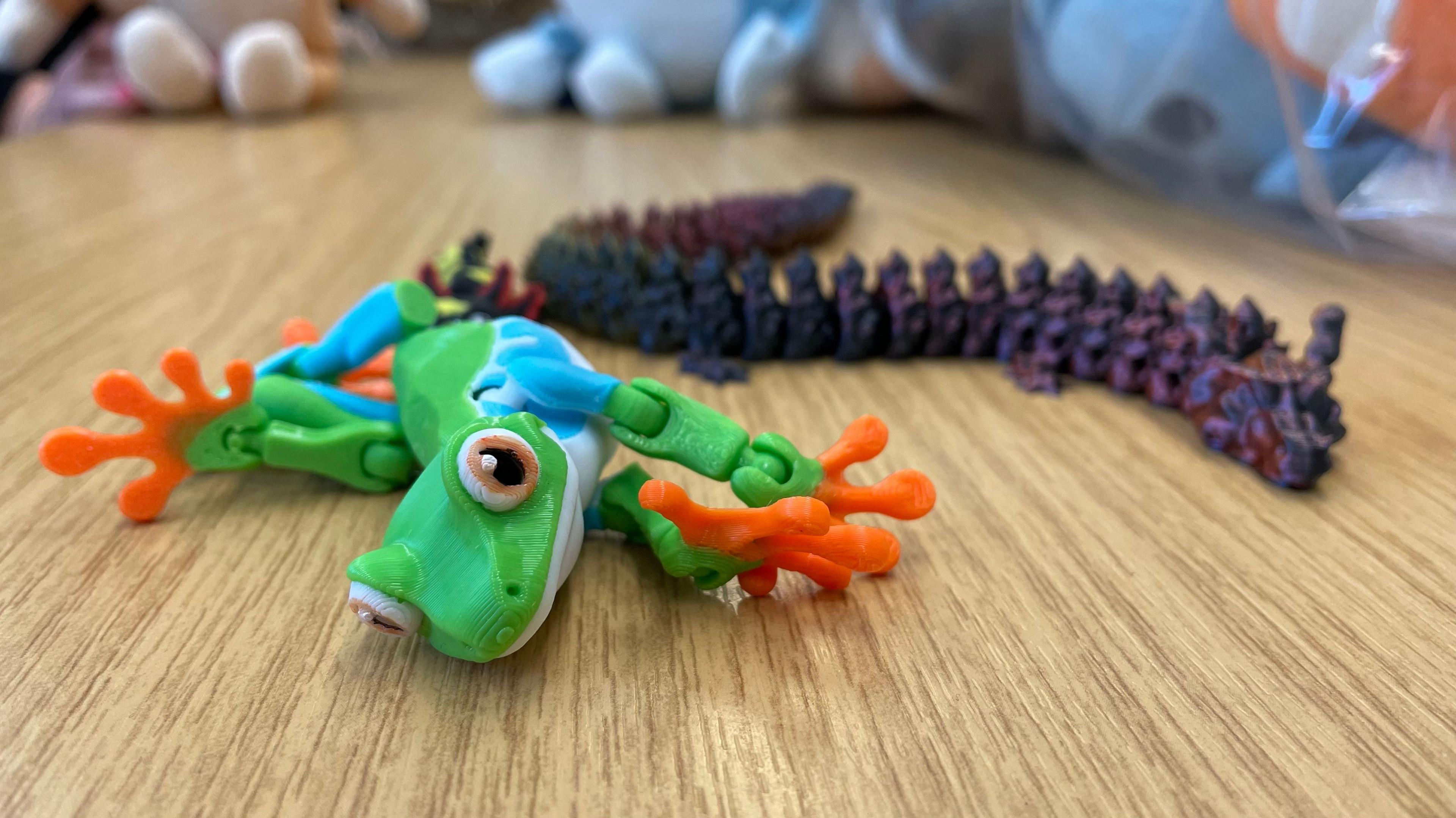 A green plastic toy frog with orange feet and flexible joints lies on a desk. It is next to a purple and red dragon toy with small parts