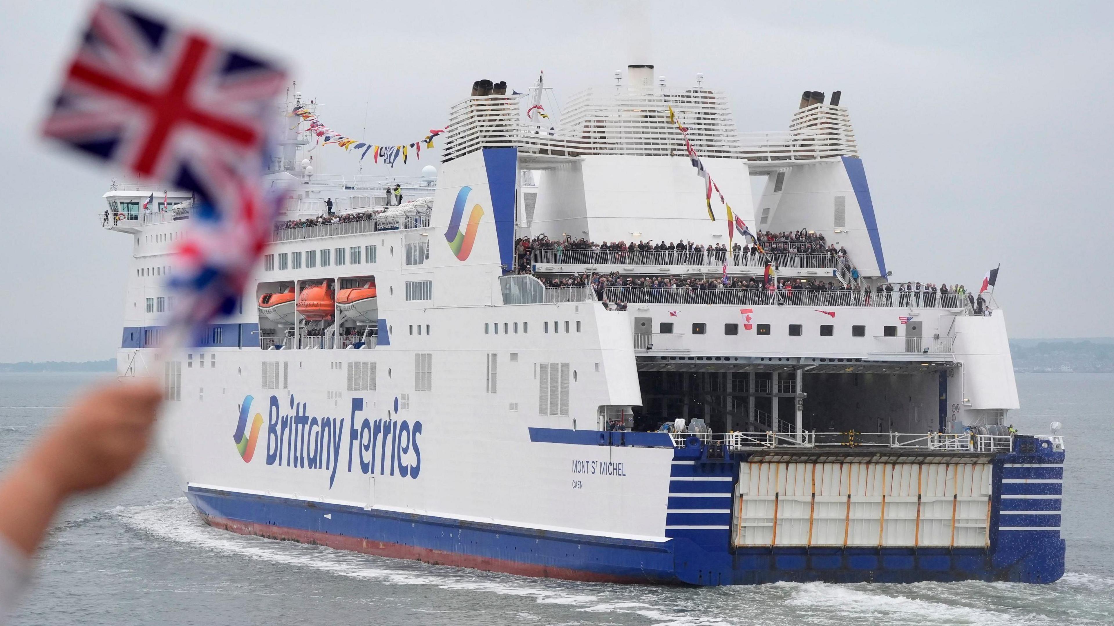 A blue and white Brittany Ferries ferry sailing away from the camera with a blurred Union Jack flag being waved in the foreground.