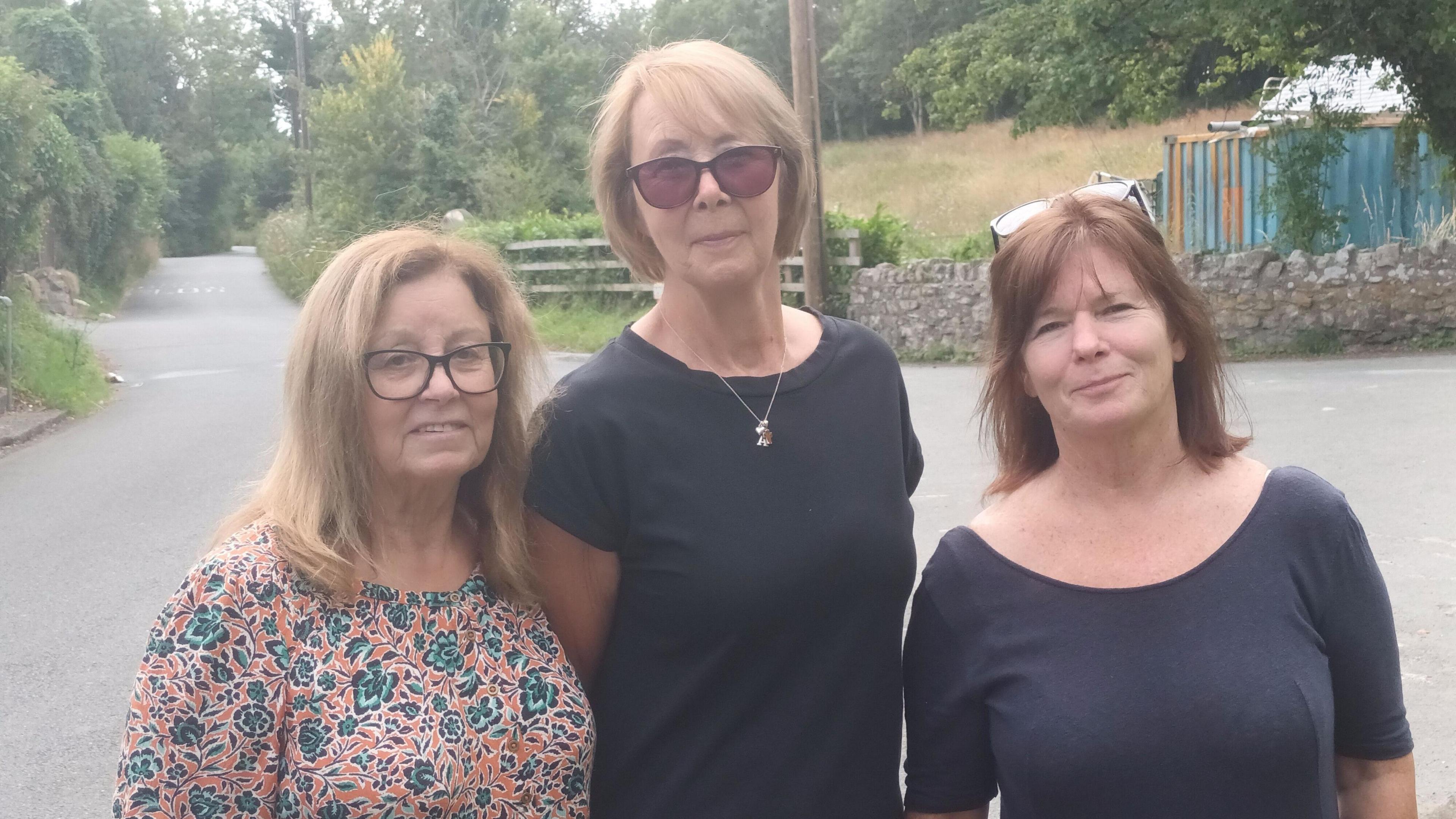 Denise Martlew, Anne Sloman and Denise Cooper pictured standing in a row by a country road with wooden fence behind. Denise is wearing an orange floral shirt with glasses and long blonde hair, Anne has short blonde hair and is wearing sunglasses, and Denise has brown hair with her glasses perched on the top of her head.