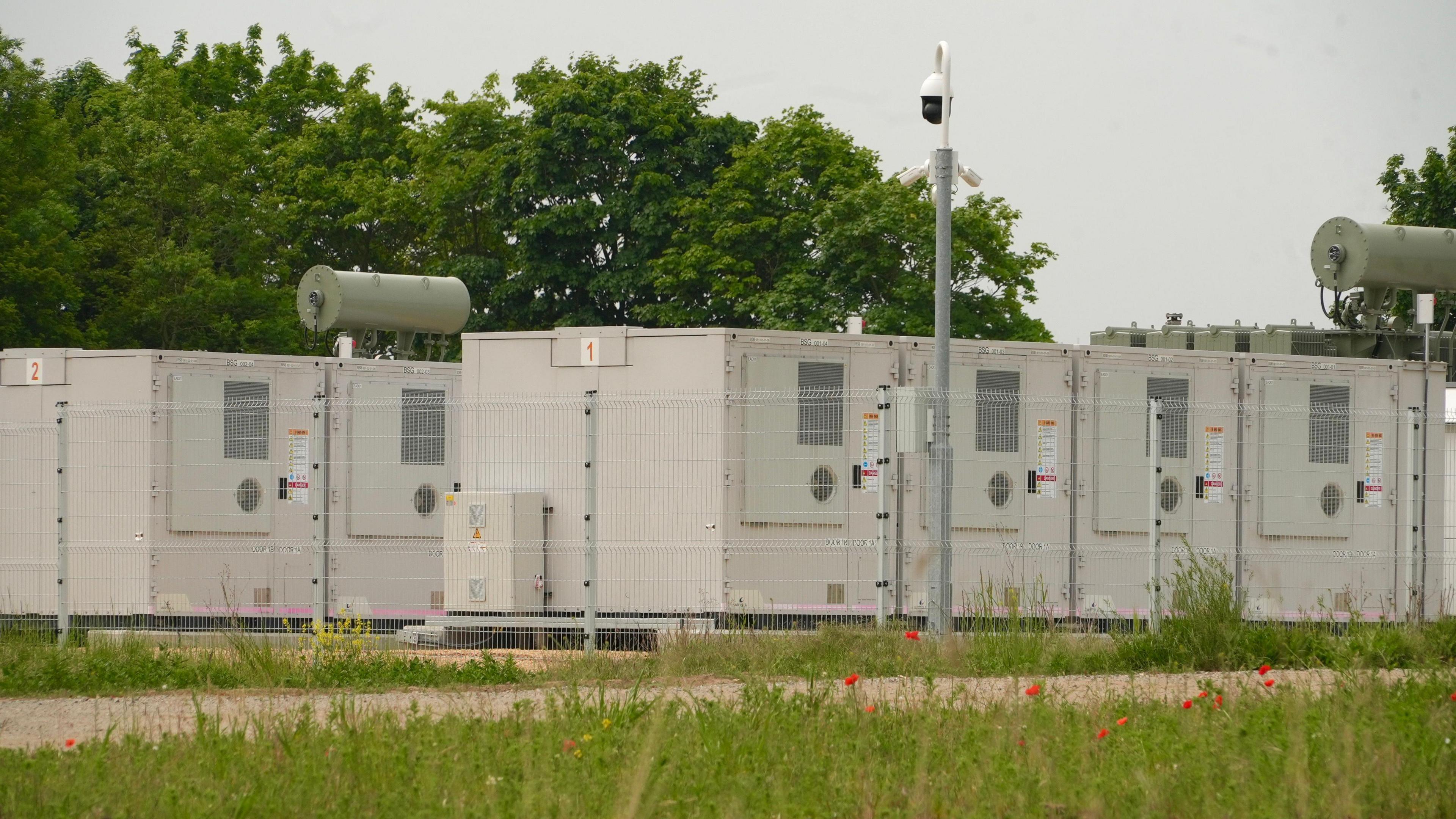 Light coloured battery storage containers, numbered 1 and 2, behind a metal fence. There are trees in the background and grass in front.