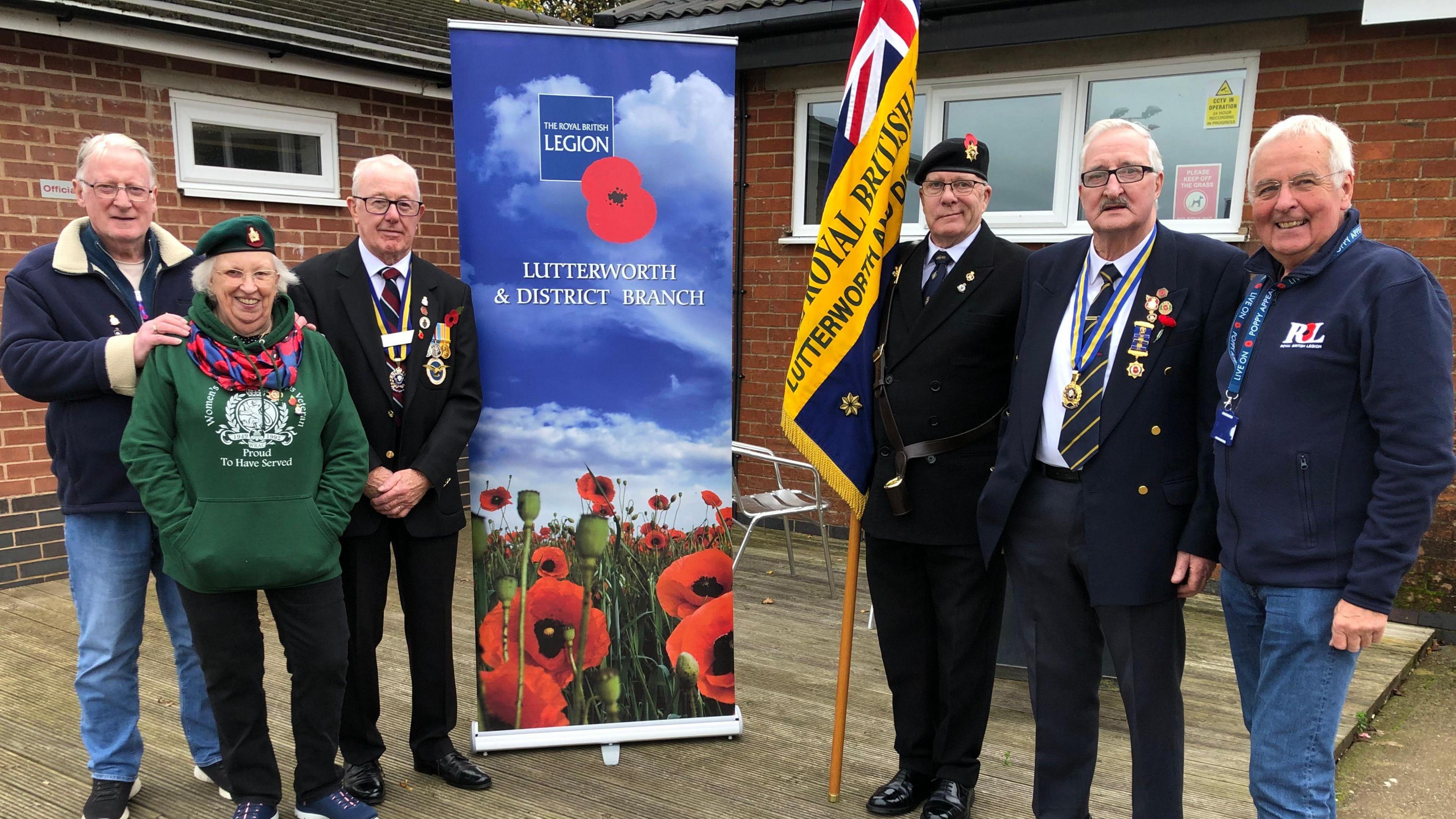 Six members of a British Legion branch pose outside a football clubhouse for a photo with a flag and a banner