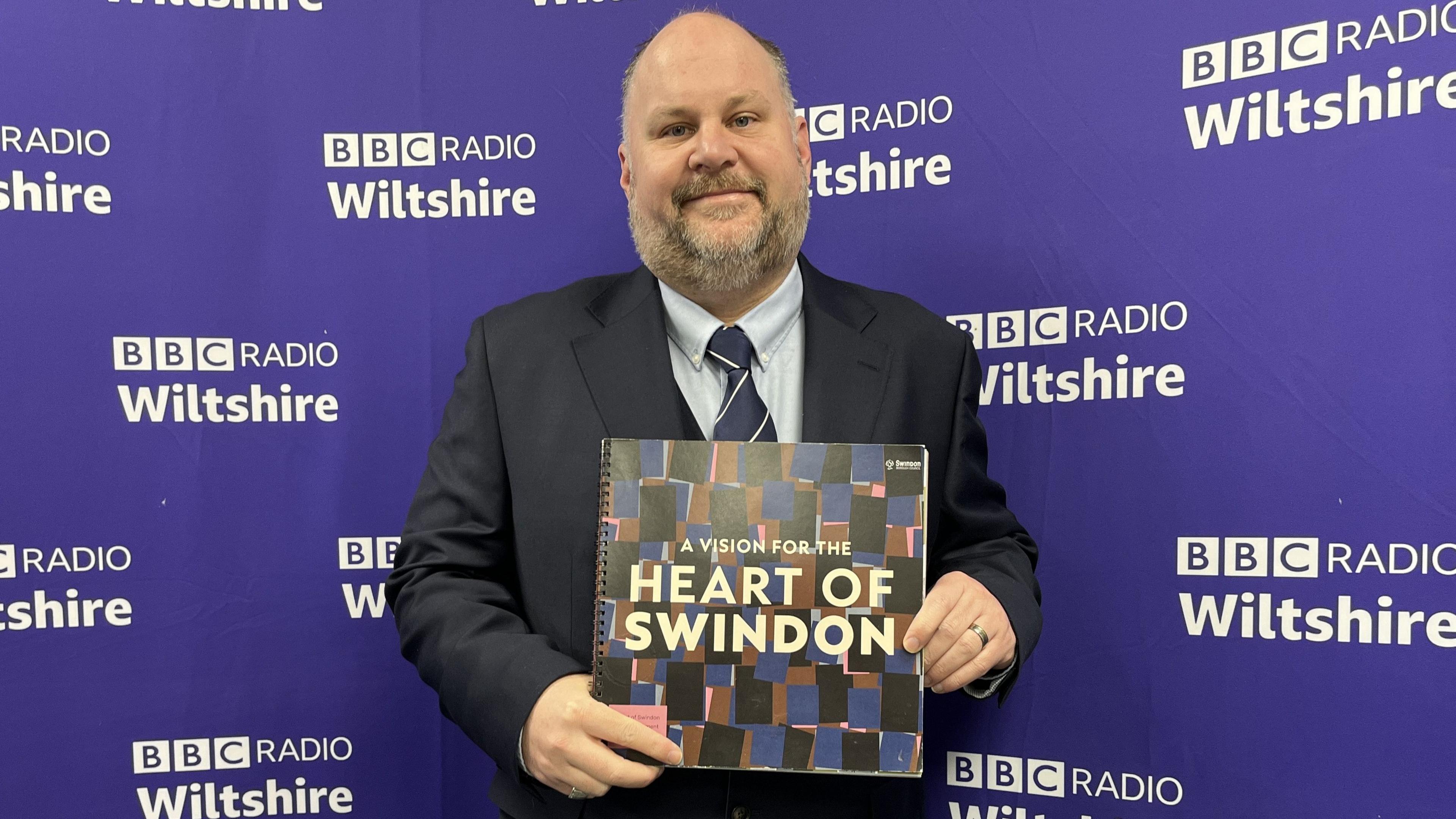 Jim Robbins, Leader of Swindon Borough Council, standing in front of a BBC Radio Wiltshire banner holding a "Heart of Swindon" brochure