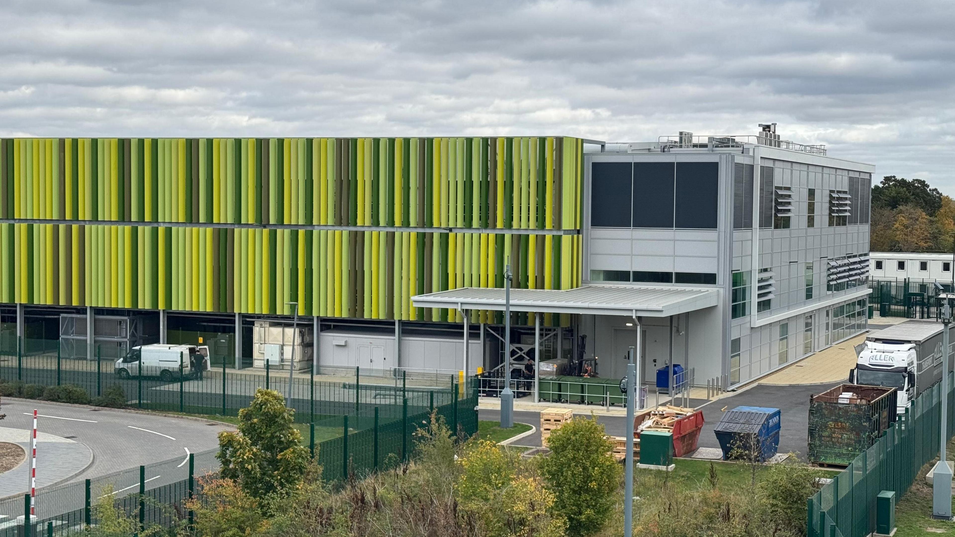 A data centre that looks like a large warehouse surrounded by tall, dark green metal fences. The building looks like it is largely covered in green stripes which run vertically down from the flat roof. The stripes are fins which help the cooling of the centre. They are in various shades of green. On the right of the warehouse are some windows which serve a number of offices inside.