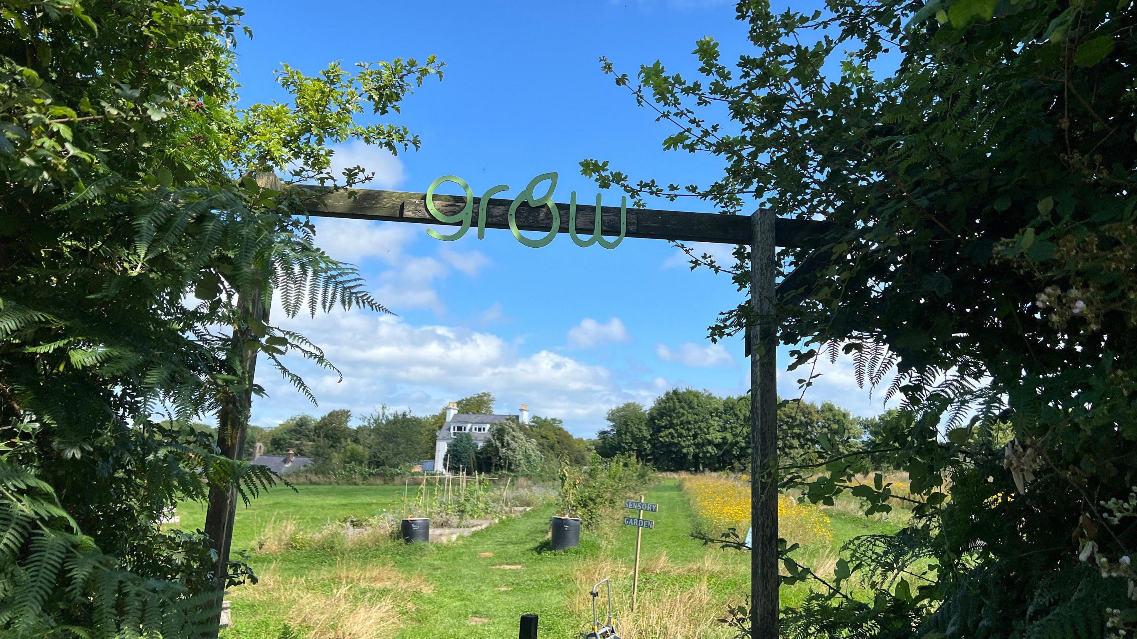 There's a wooden arch over the entrance to a field where fruit and vegetables are being grown. The word Grow is on the arch with the letter o represented as an apple