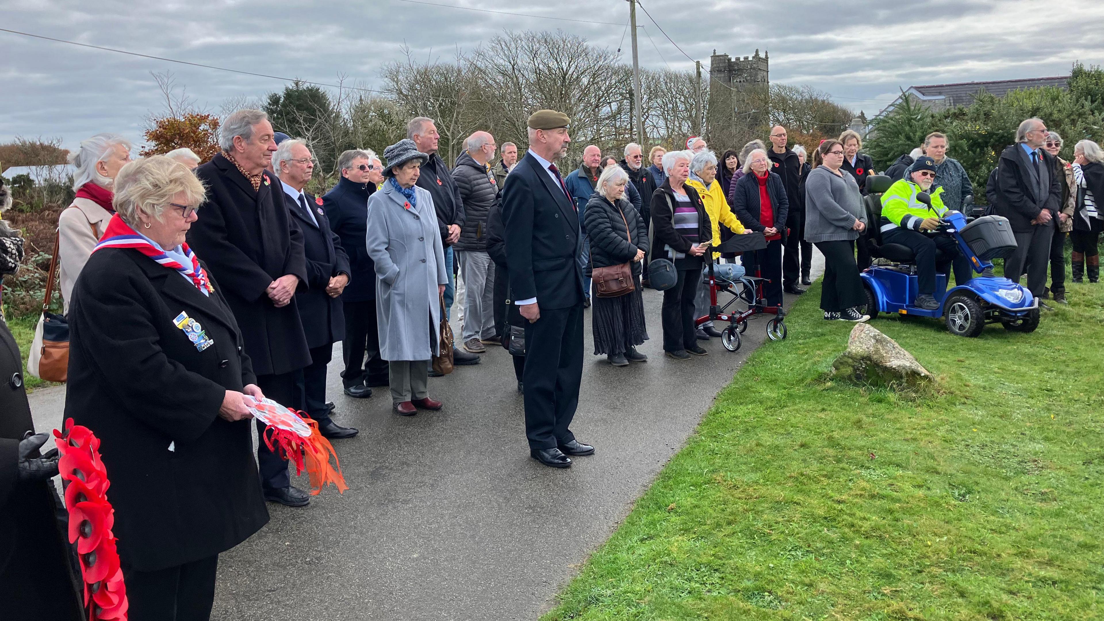 A group of people gathered at the memorial service in St Breward. There is a poppy wreath on the left and people looking towards the monument. The people are stood on the pavement with some standing on the grass on the right.