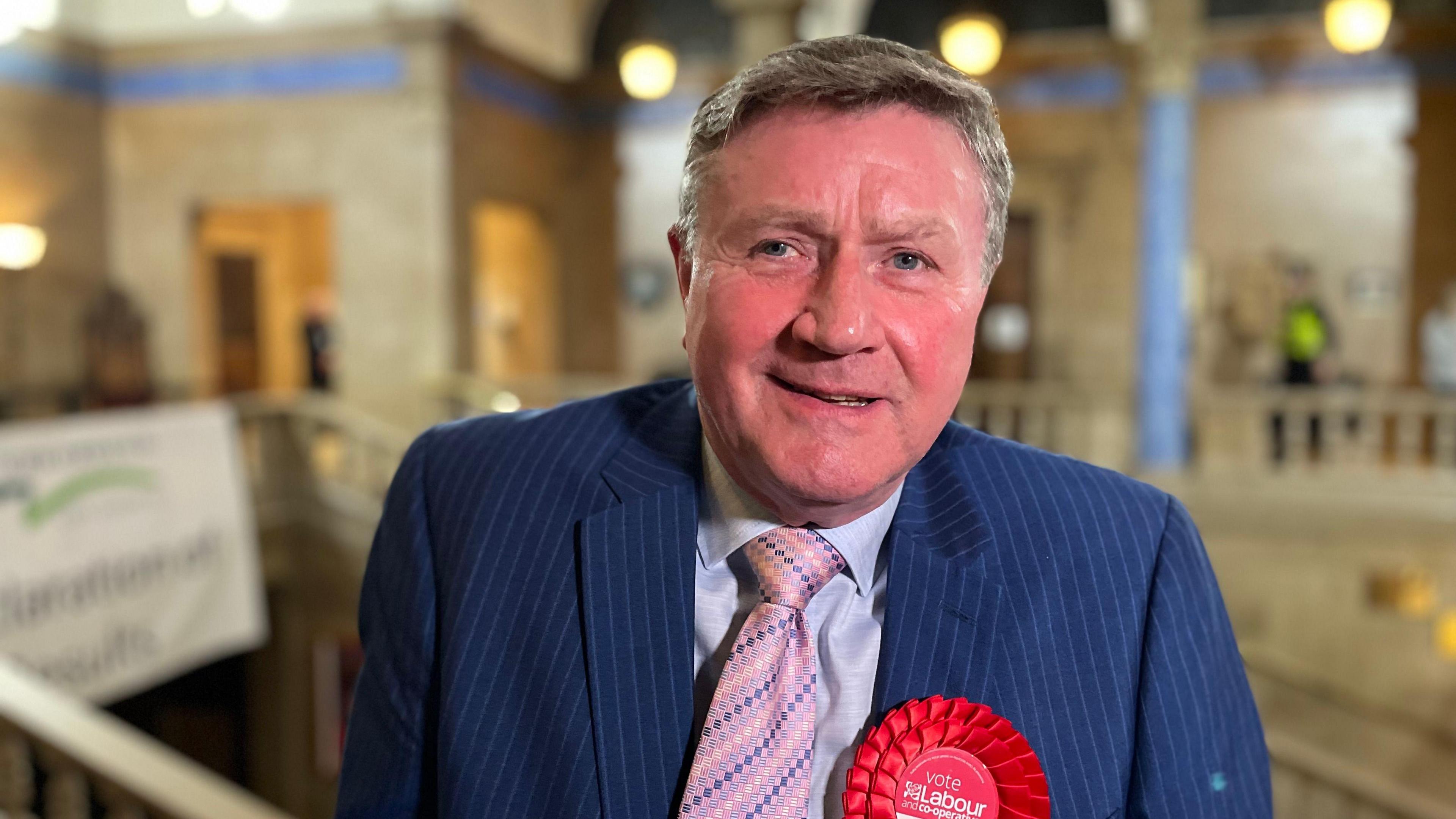 Dennis Jones wearing a navy pinstriped blazer, light coloured shirt and pink tie as well as a red Labour rosette. He is standing in Peterborough Town Hall and looking at the camera.