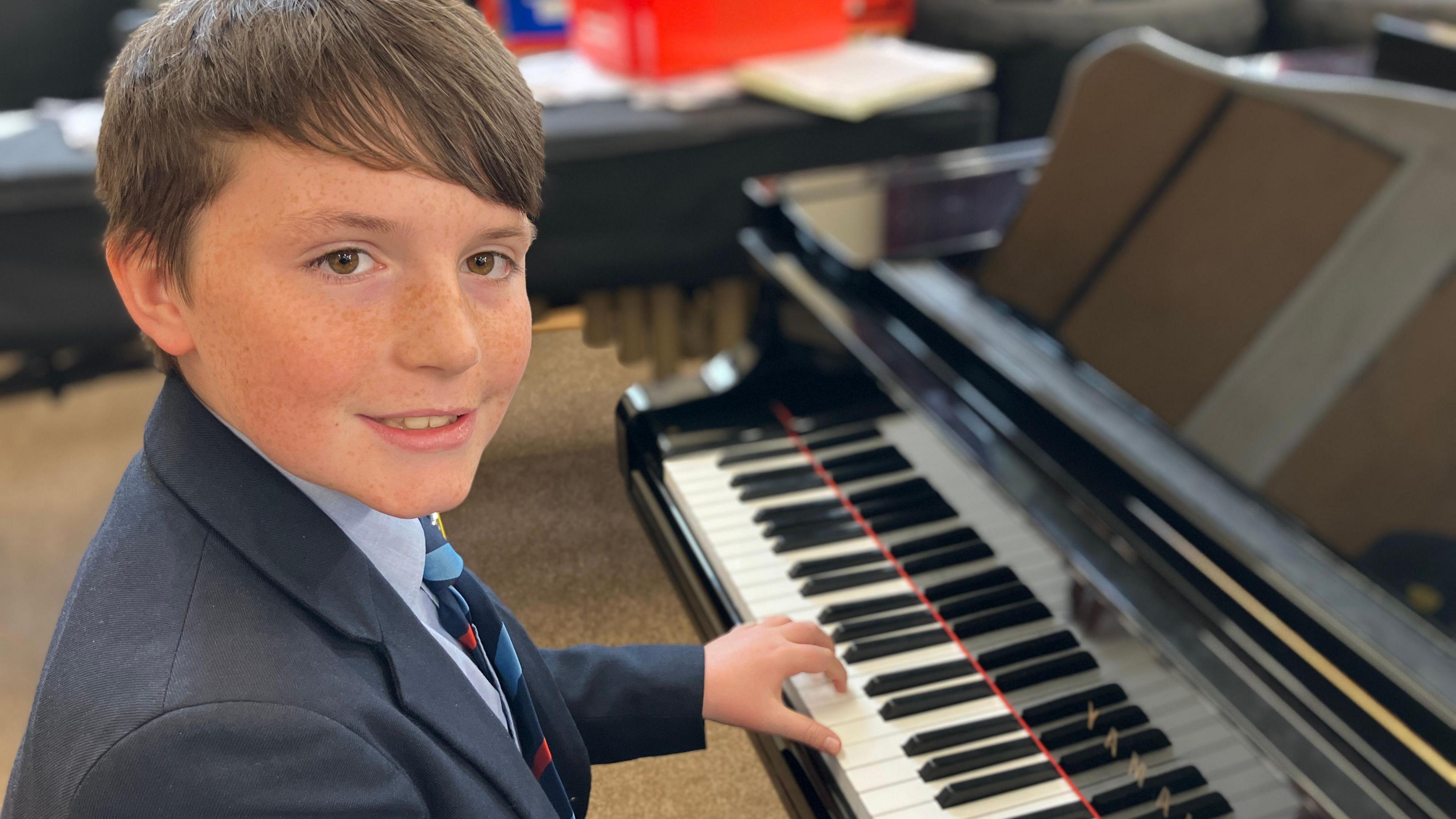 River sat at a piano wearing a navy jacket and blue shirt with his blue and red school tie. He has short brown hair and brown eyes.