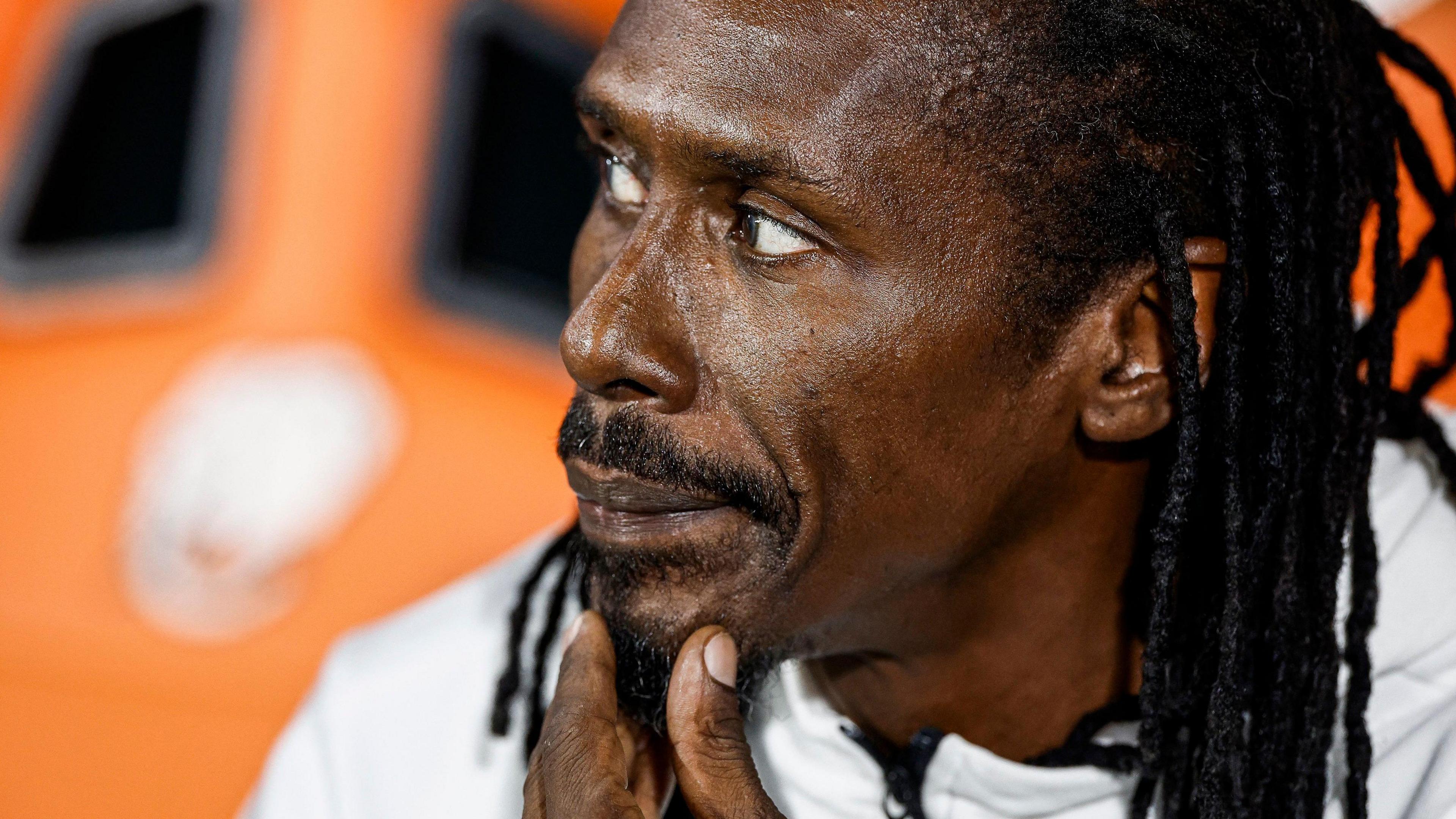 Aliou Cisse, with his back hair braided and a goatee beard, holds his left hand to his chin as he looks to his right