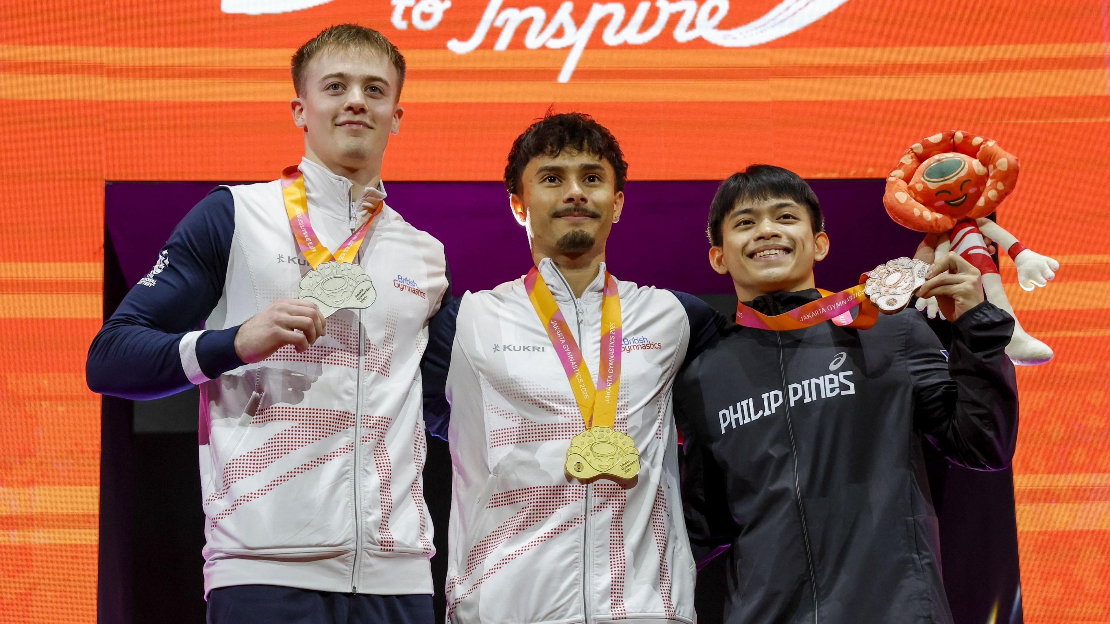 Luke Whitehouse (left), Jake Jarman (centre) and Carlos Yulo (right) celebrate their win on a podium with medals