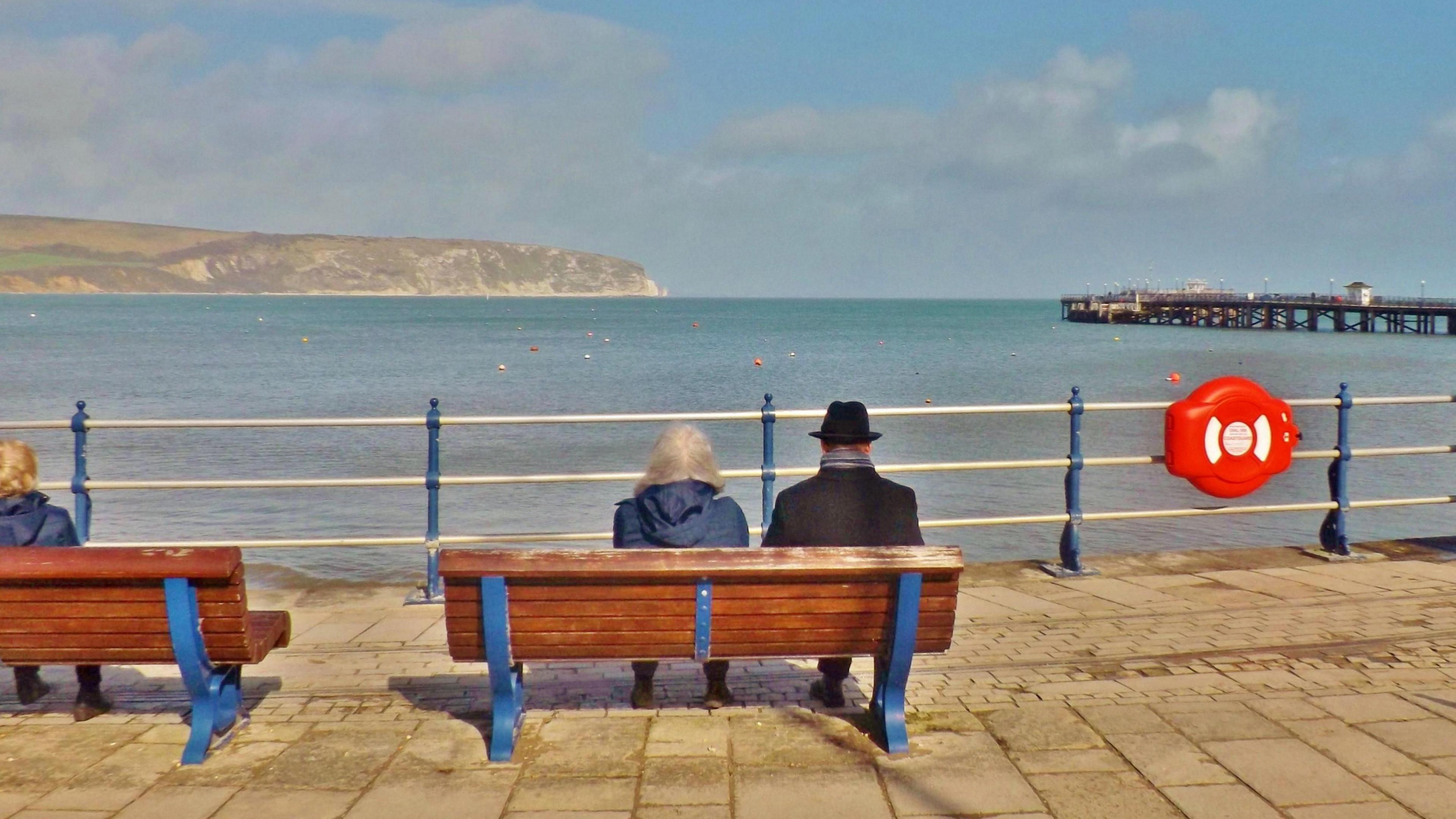 Two people sit on a bench overlooking the sea on a sunny day. One is wearing a black hat and coat, the other has long grey hair and is wearing a blue coat. In front of them are white railings.  In the distance you can see a wooden piece and a cliff.