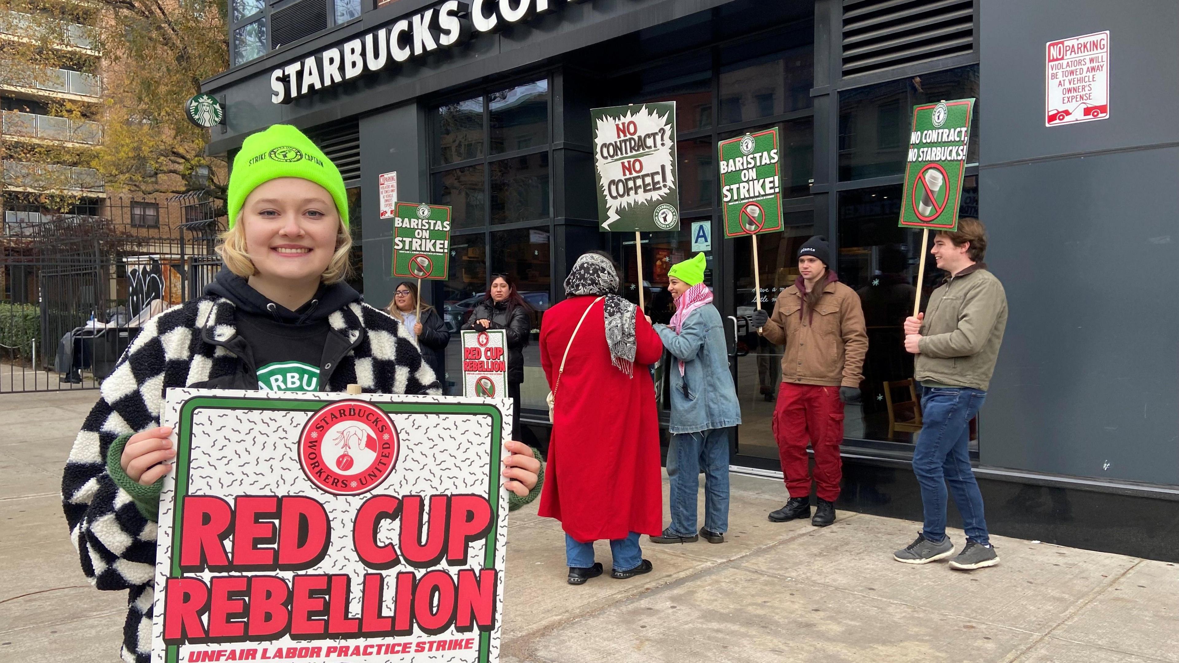 A woman wearing a neon beanie holds a sign reading "Red Cup Rebellion", standing in front of a Starbucks Coffee store alongside other people holding signs.