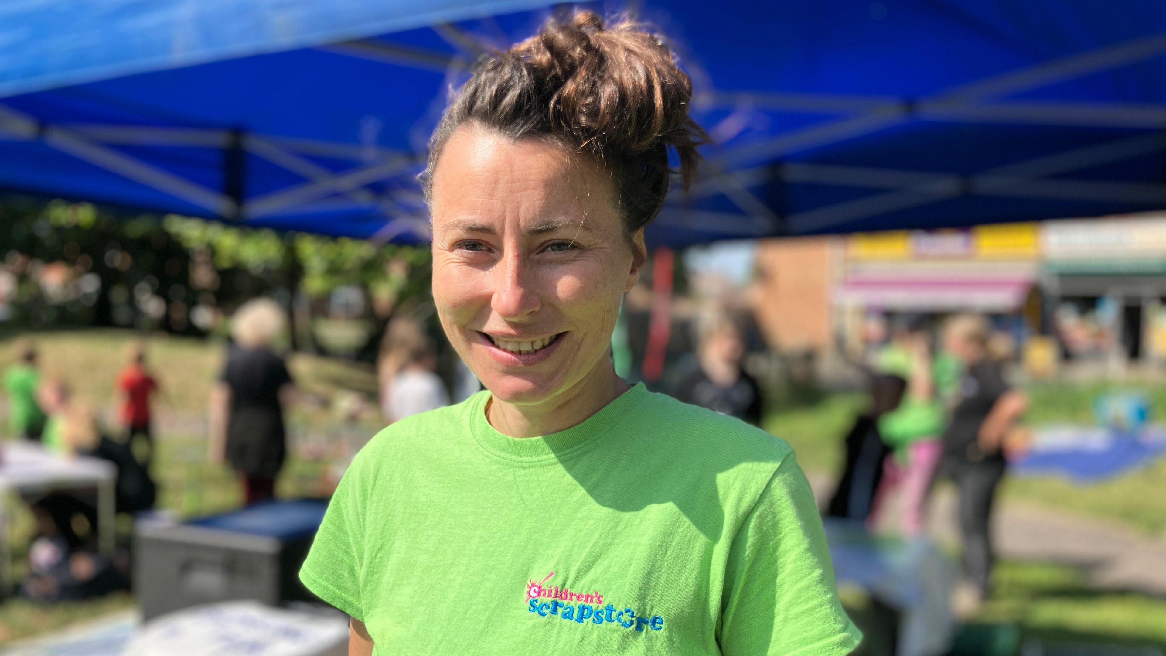 Alys Scratchley with her brown hair tied up in a bun, wearing a bright green t-shirt that says 'Children's Scrapstore' on it. She is standing in front of a blue cabana tent where there are tables set up, and children playing together in the background