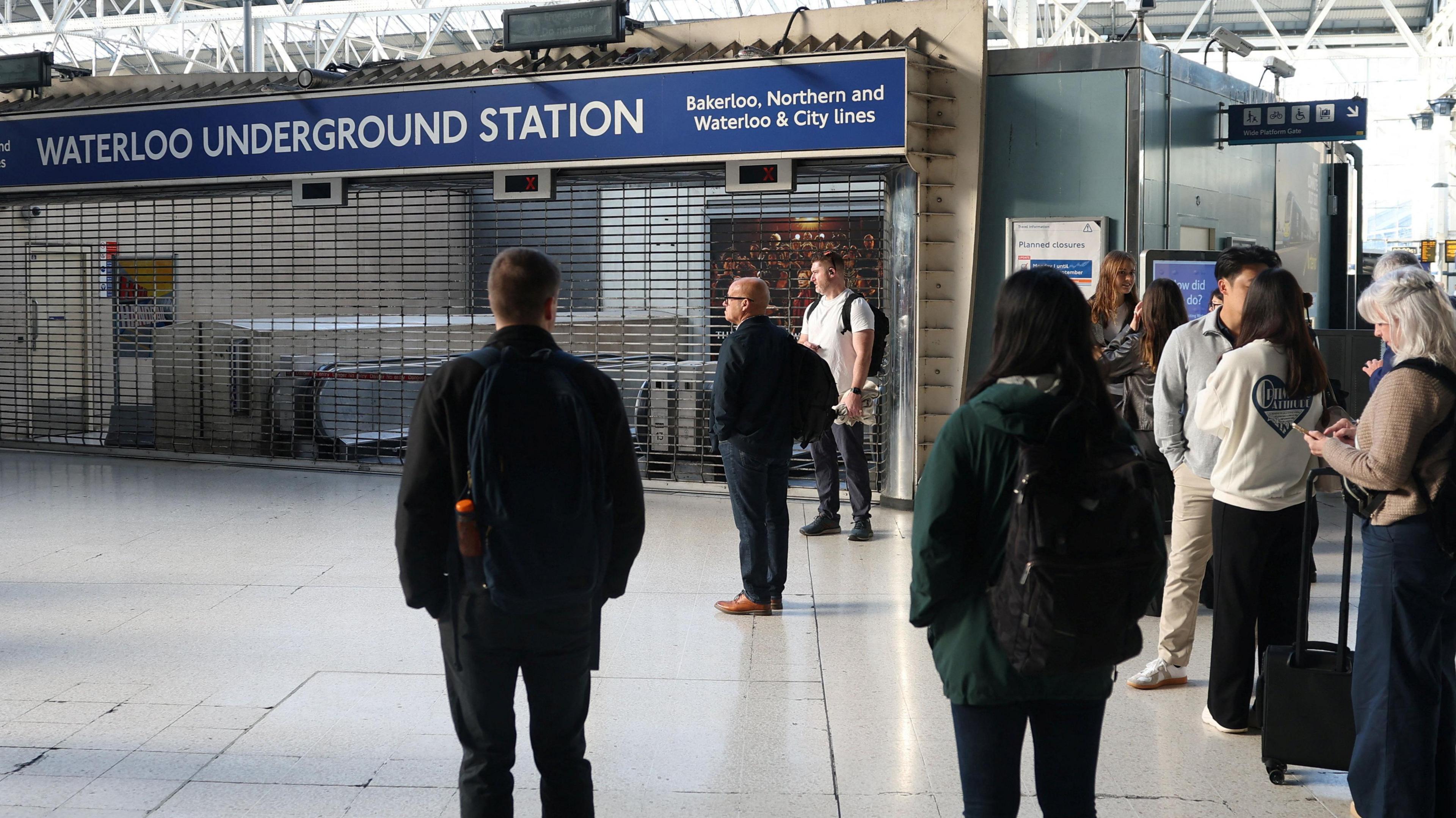 Commuters stand outside the closed entrance to Waterloo Underground Station, where shutters are pulled down.