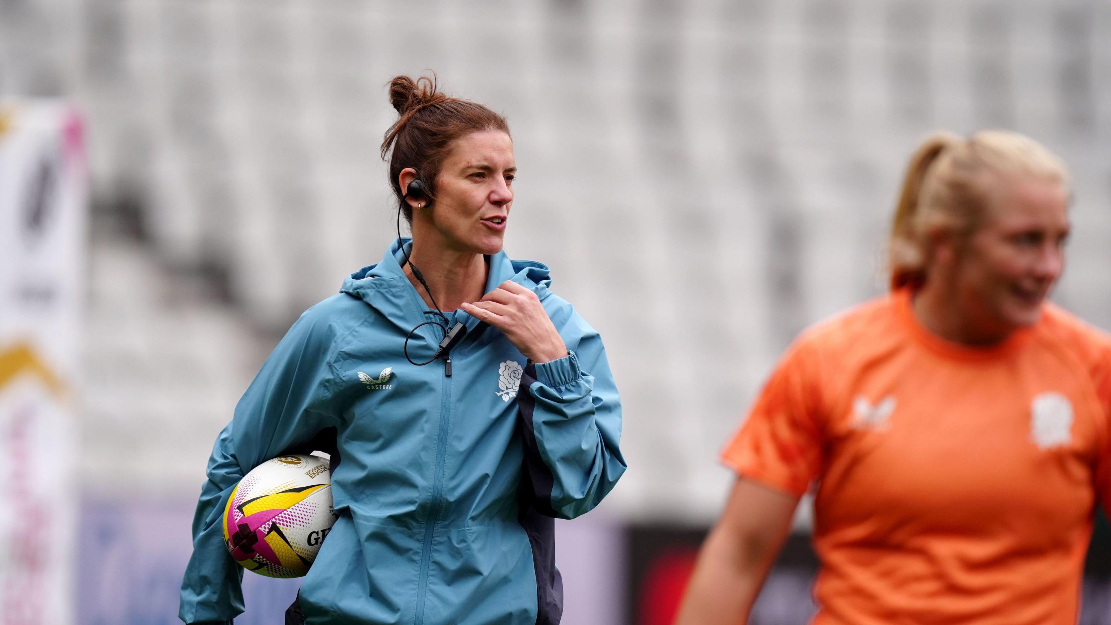 Sarah Hunter during an England training session. She is wearing her blue England coach's kit and is holding a white rugby ball under her arm. An England player stand to the right in her orange training kit.