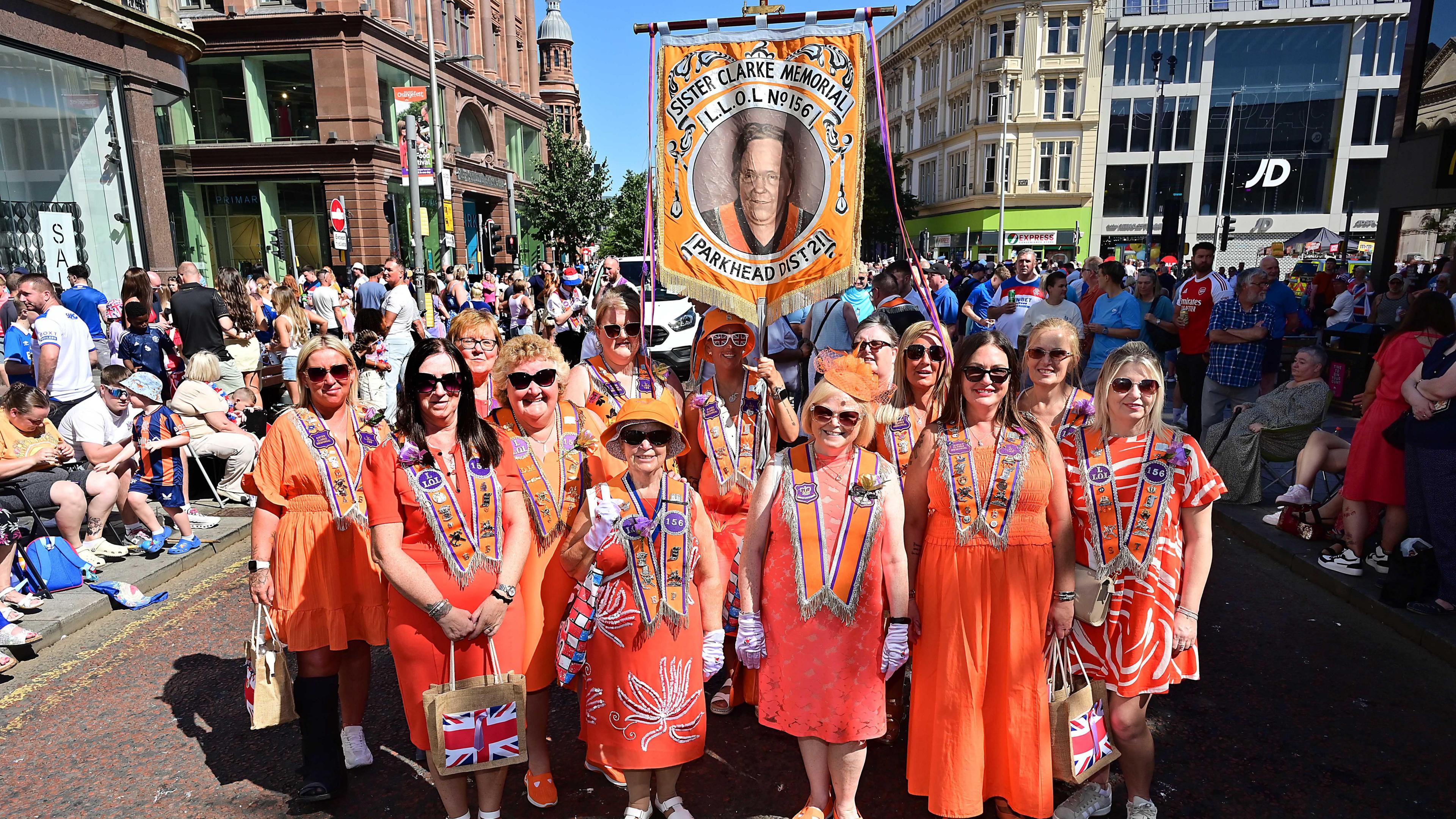 A group of women in orange dresses and sashes wearing sunglasses as they pose on a street in Belfast and hundreds of spectators are behind them. The women are holding a banner that says "Sister Clarke Memorial L.L.O.L No. 156 Parkhead Dist 2". The sun is shining on the women.