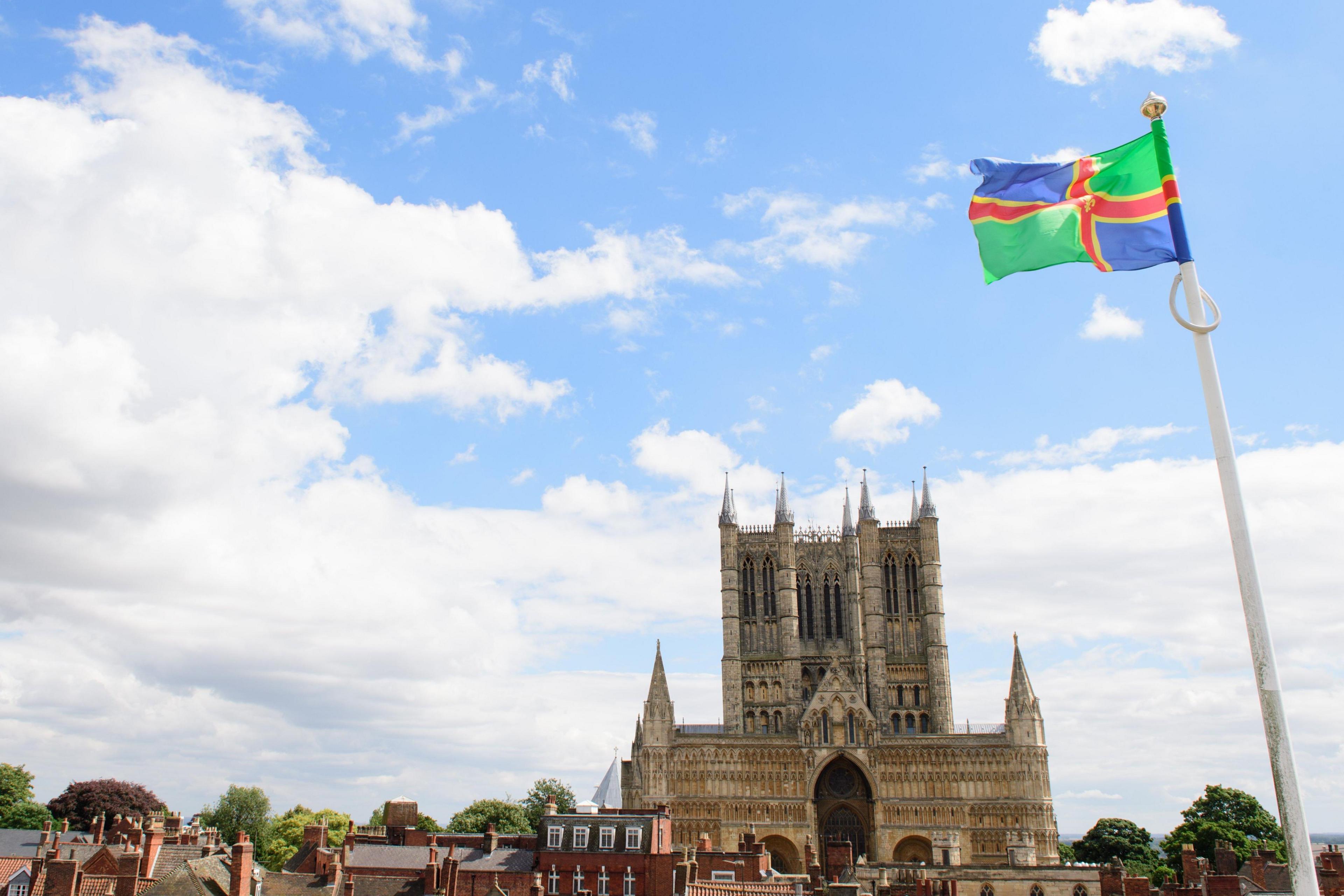 Lincoln Cathedral, a historic Gothic-style building stands prominently on the skyline with tall spires and intricate stonework. The rooftops of nearby buildings and a flagpole displaying a colourful flag with green, blue, red, and yellow stripes are in the foreground. The sky is blue with patchy cloud.