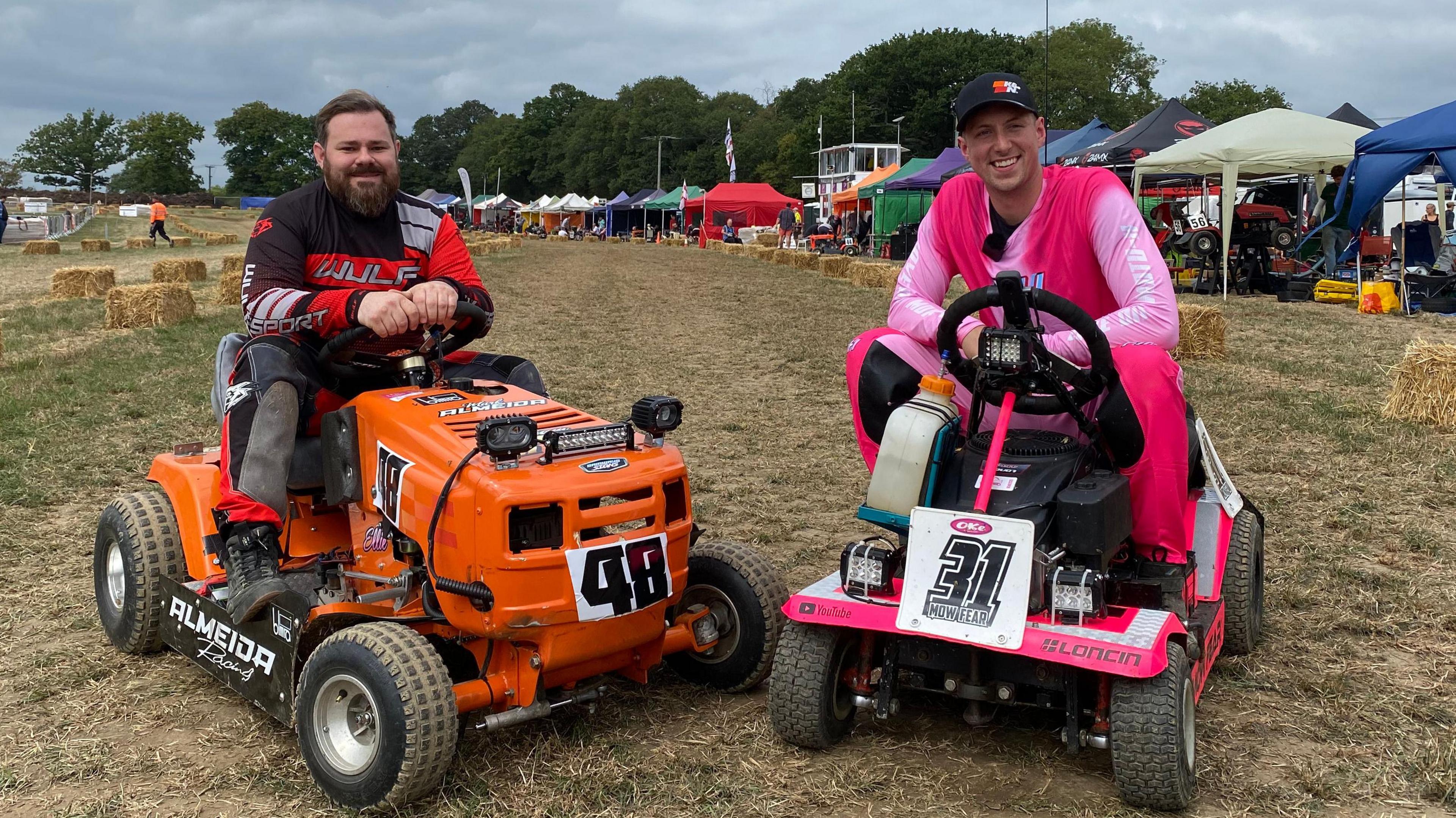Two men are sat on lawn mowers with racing colours and numbers, smiling at the camera