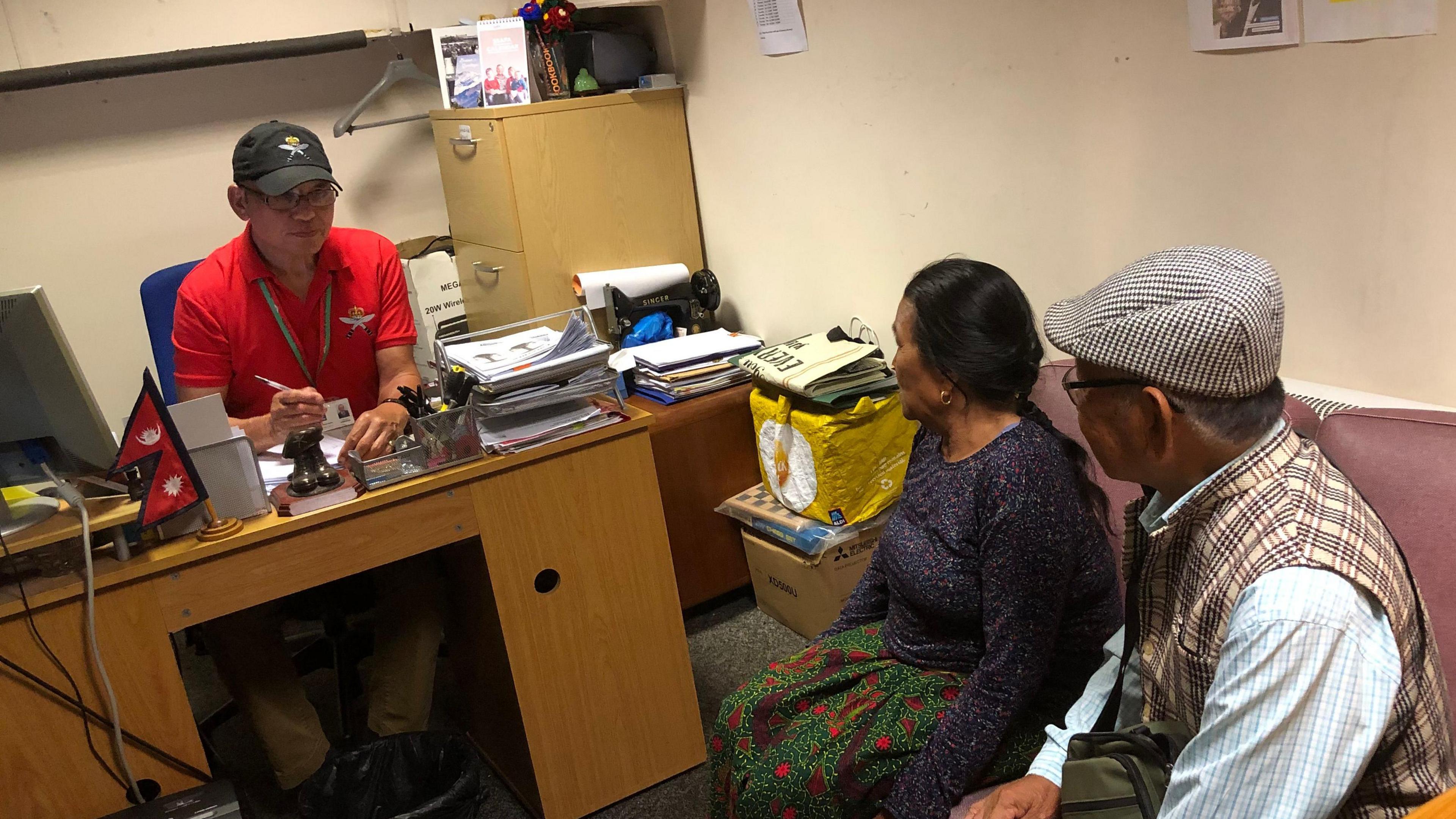 Chandra Budhathoki in a red polo shirt and black baseball cap. He grasps a pen and is writing on some paper as he sits behind a desk. In front of him is an elderly many and woman who are looking to him for support and talking with him.