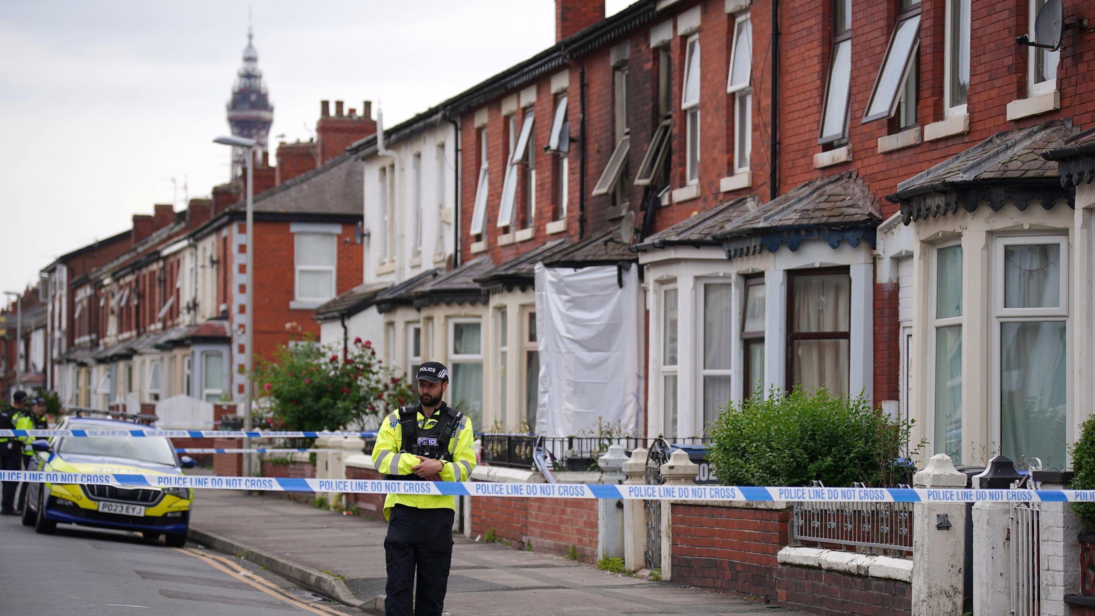 A row of terraced houses with a blackened front of one of them where a fire was. Blackpool Tower can be seen in the background. A police officer in uniform walks near a cordon, with a police car behind.