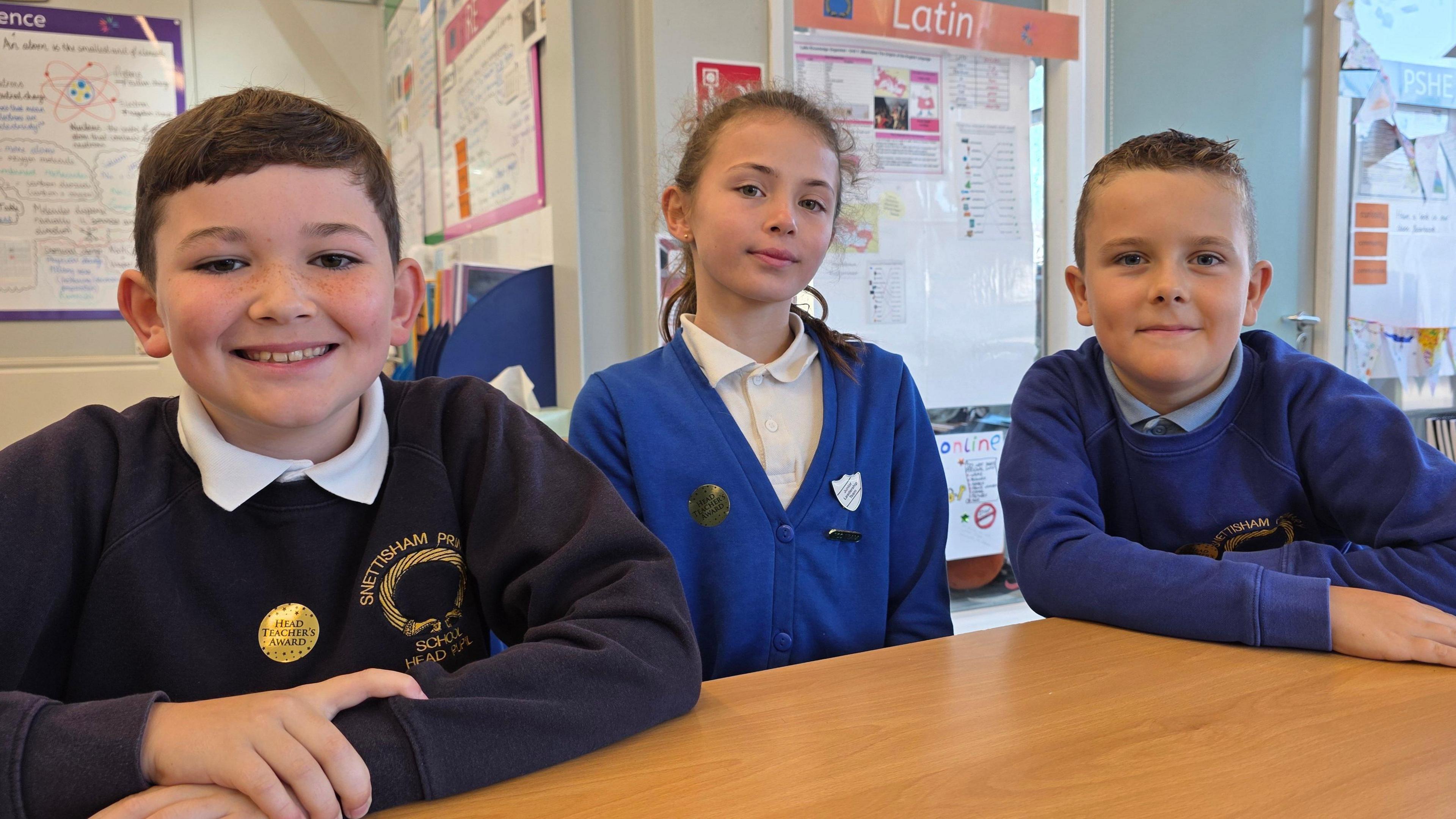 We can see three children in a classroom. On the left is a boy wearing a dark blue jumper on top of a white polar neck. In the middle is a girl wearing a blue cardigan over a white shirt. And on the right is another boy wearing a blue jumper over a blue polar neck.