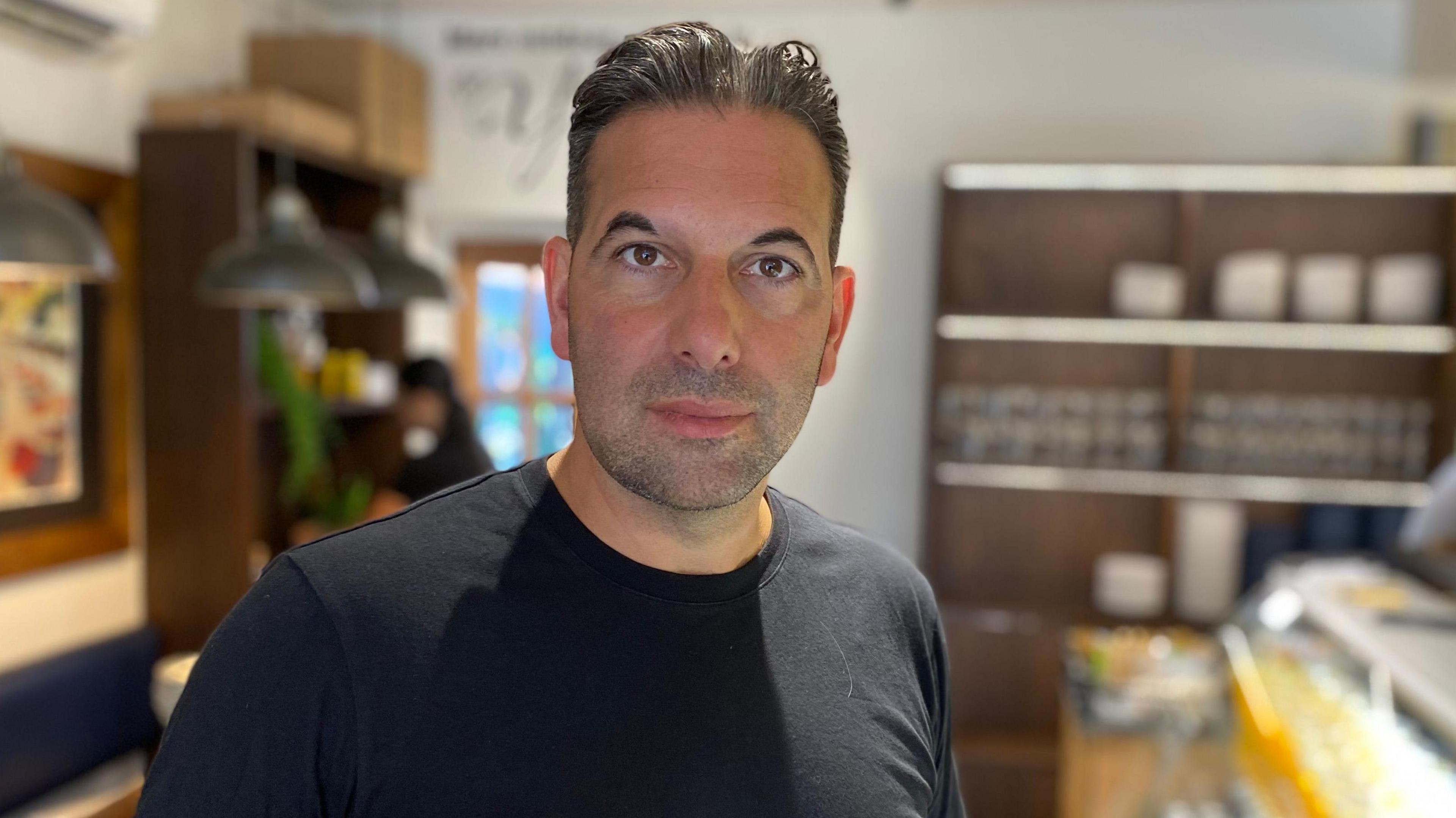 Deli owner John Hyman inside shop in West Bridgford, Nottinghamshire, wearing a dark t-shirt and looking at the camera.