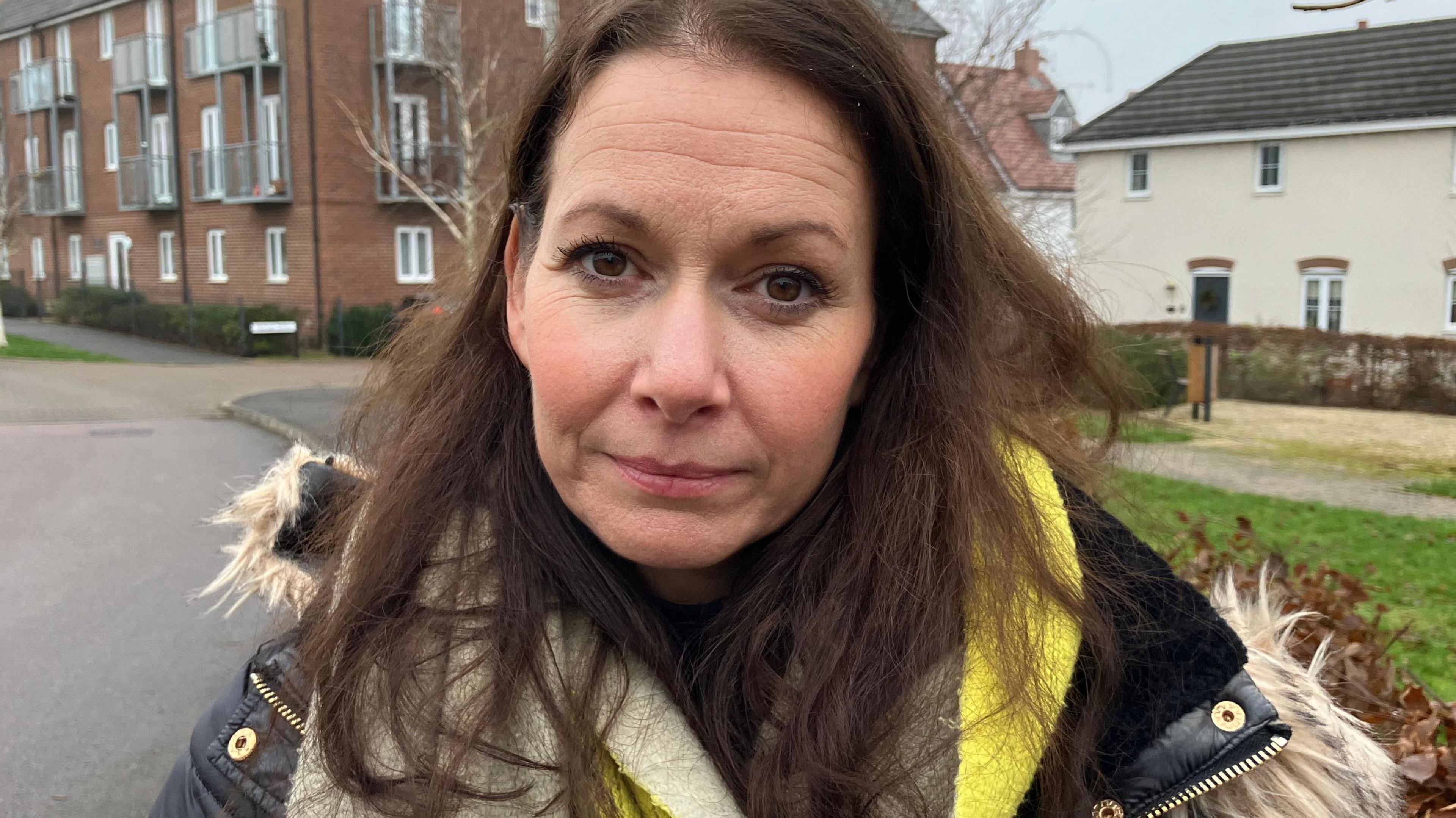 Sophie Pritchard stands on the estate. She is looking at the camera with a solemn expression. She is wearing a padded blue coat, bright green, beige and brown scarf, and has long brown hair and brown eyes. 