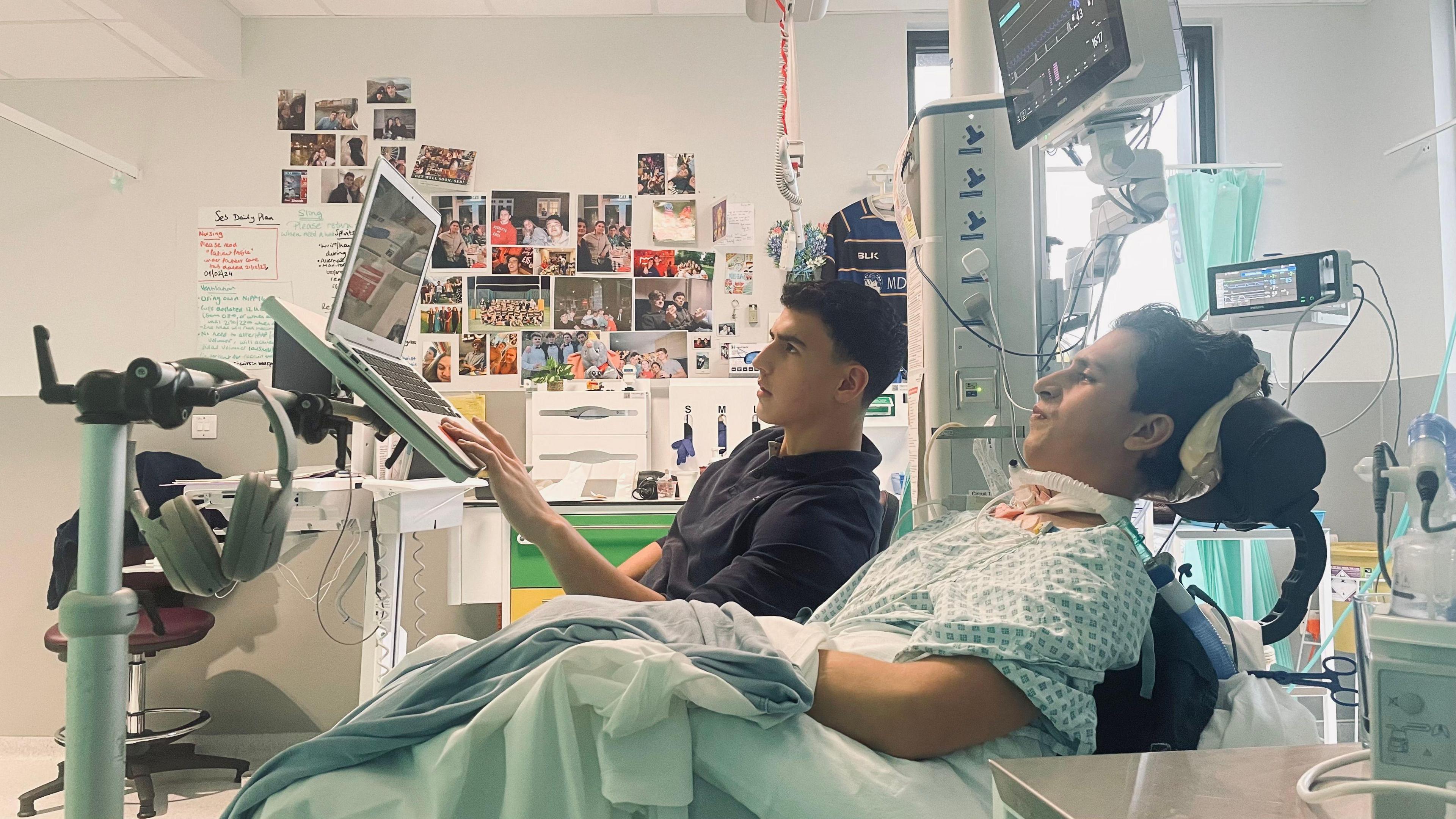 Seb in his hospital bed, with lots of equiptment atached to him. His brother is sat next to him and scrolling on a laptop propped up in front of Seb. In the background is a wall covered with images of Seb with his friends and family.