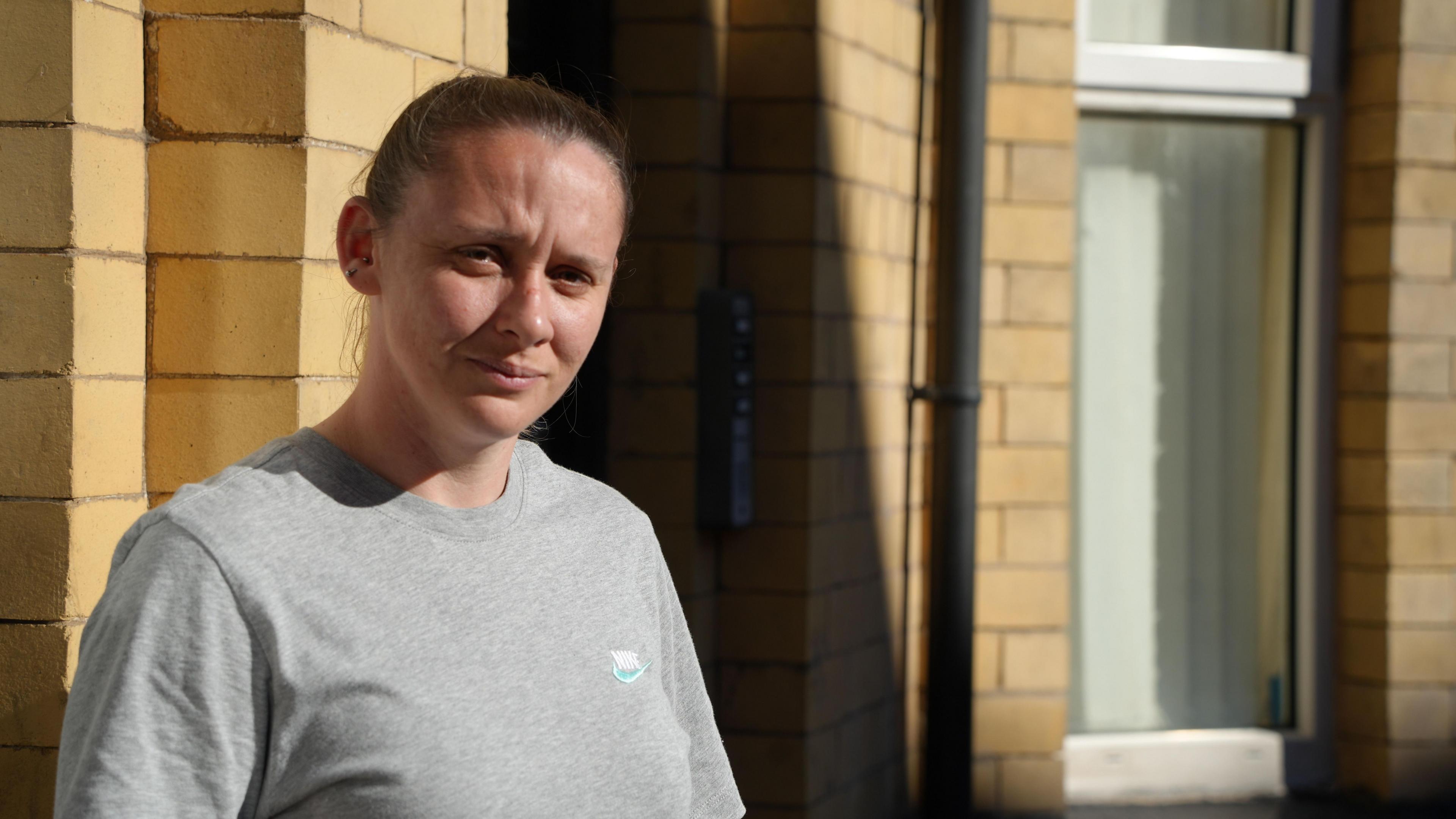 Elissa Hubbard wearing a grey Nike T-shirt and looking at the camera. She is standing in front of her home which is yellow brick and a window is visible in the background