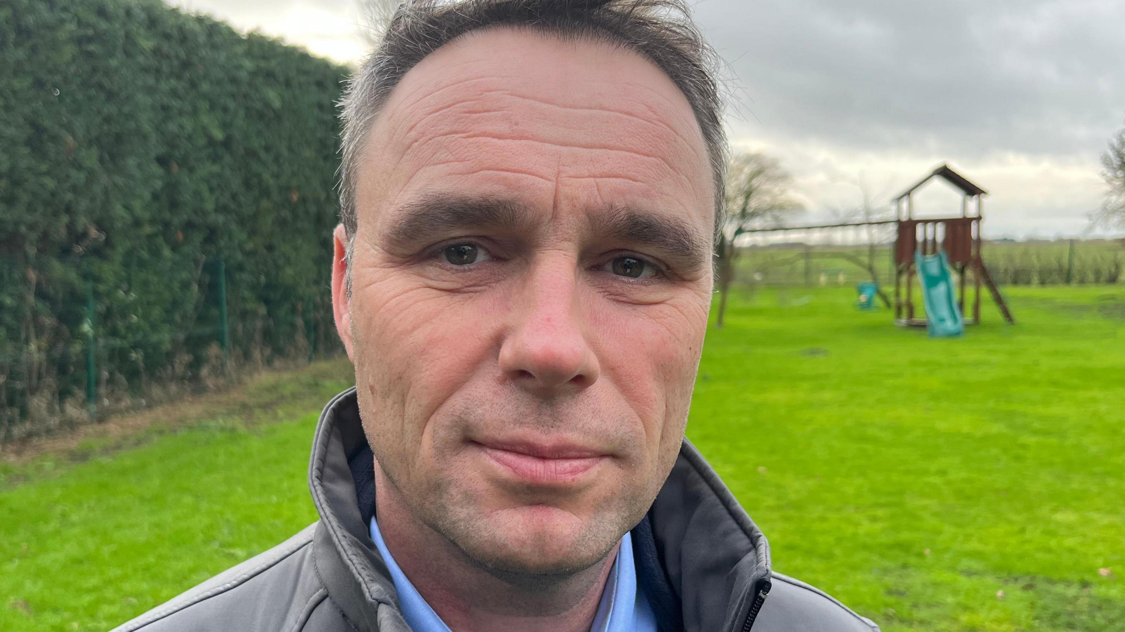 Mathew Latta is stood on the well-kept green grass in his back garden. He looks concerned. He has green eyes, grey hair and a wears a blue shirt and a grey gilet.