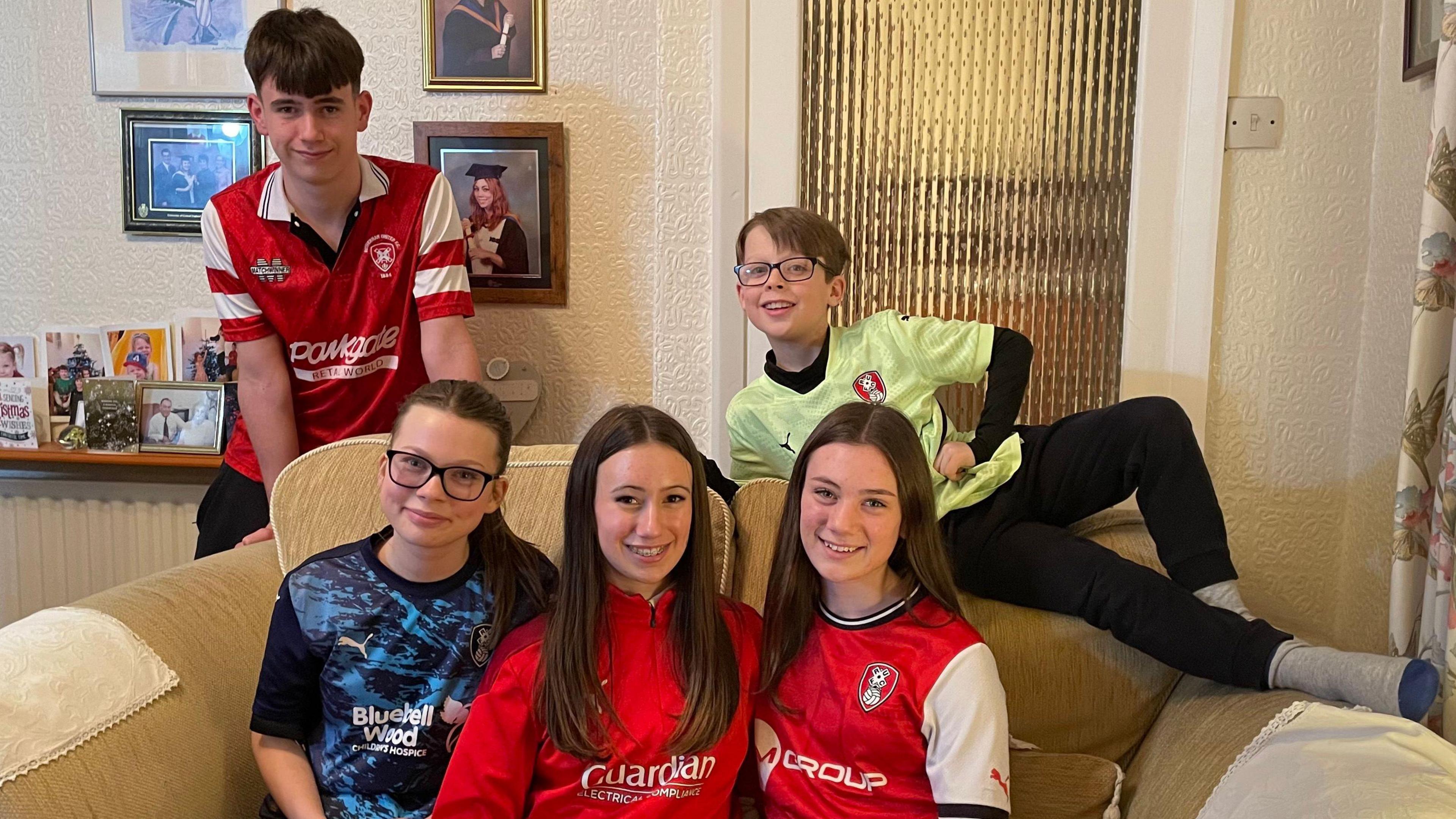 Five Millers-branded children. The three girls are sat together on a sofa. The eldest, Charlie, stands behind them. The youngest, Sam, is laid across the top of the cushions with his hand on his hip.