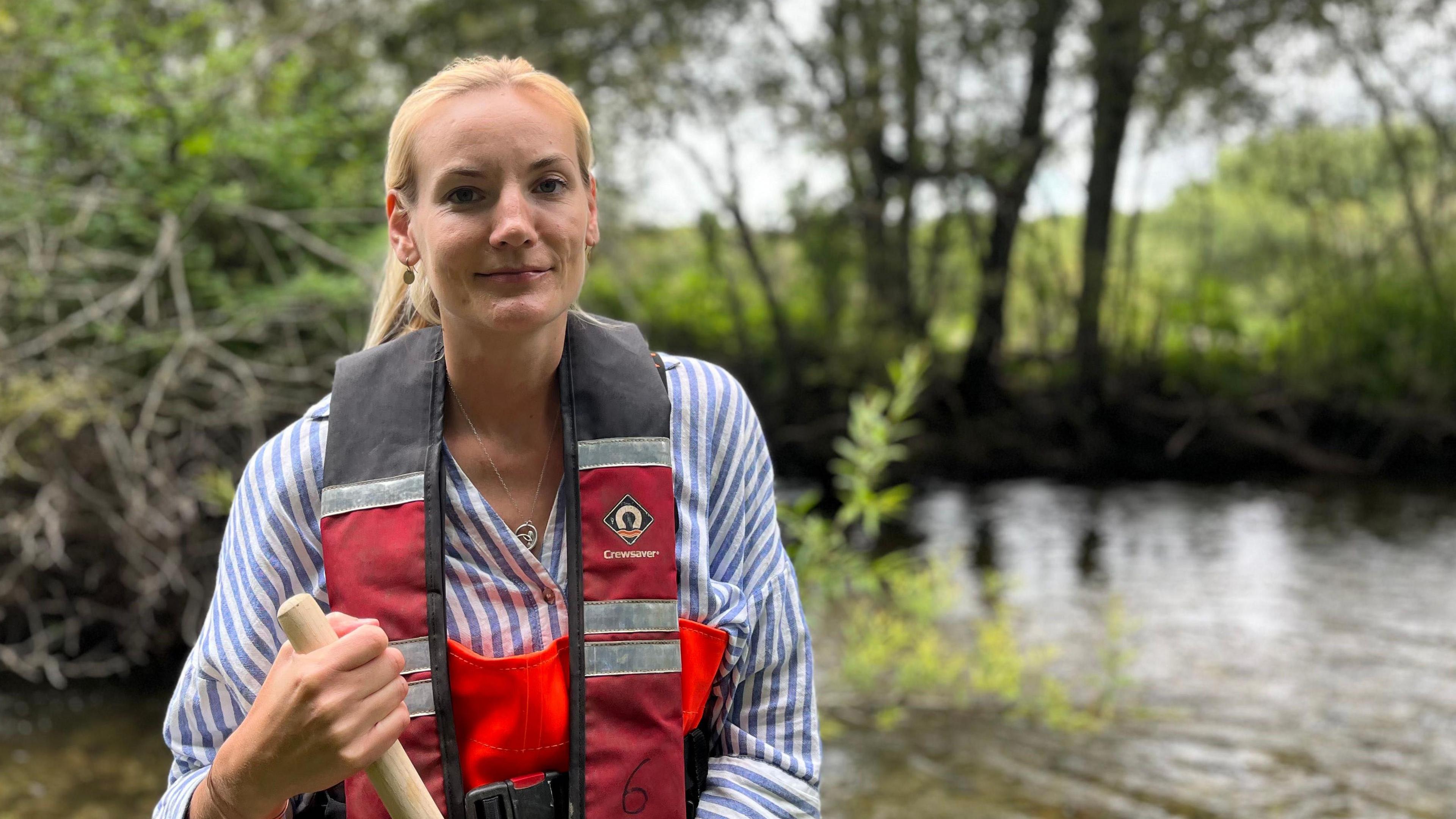 Alice smiles at the camera. She has her blonde hair tied up and wears orange river waders, a blue and red life jacket, and a blue striped shirt underneath. She holds a wooden pole and stands in front of a river, blurred in the background.