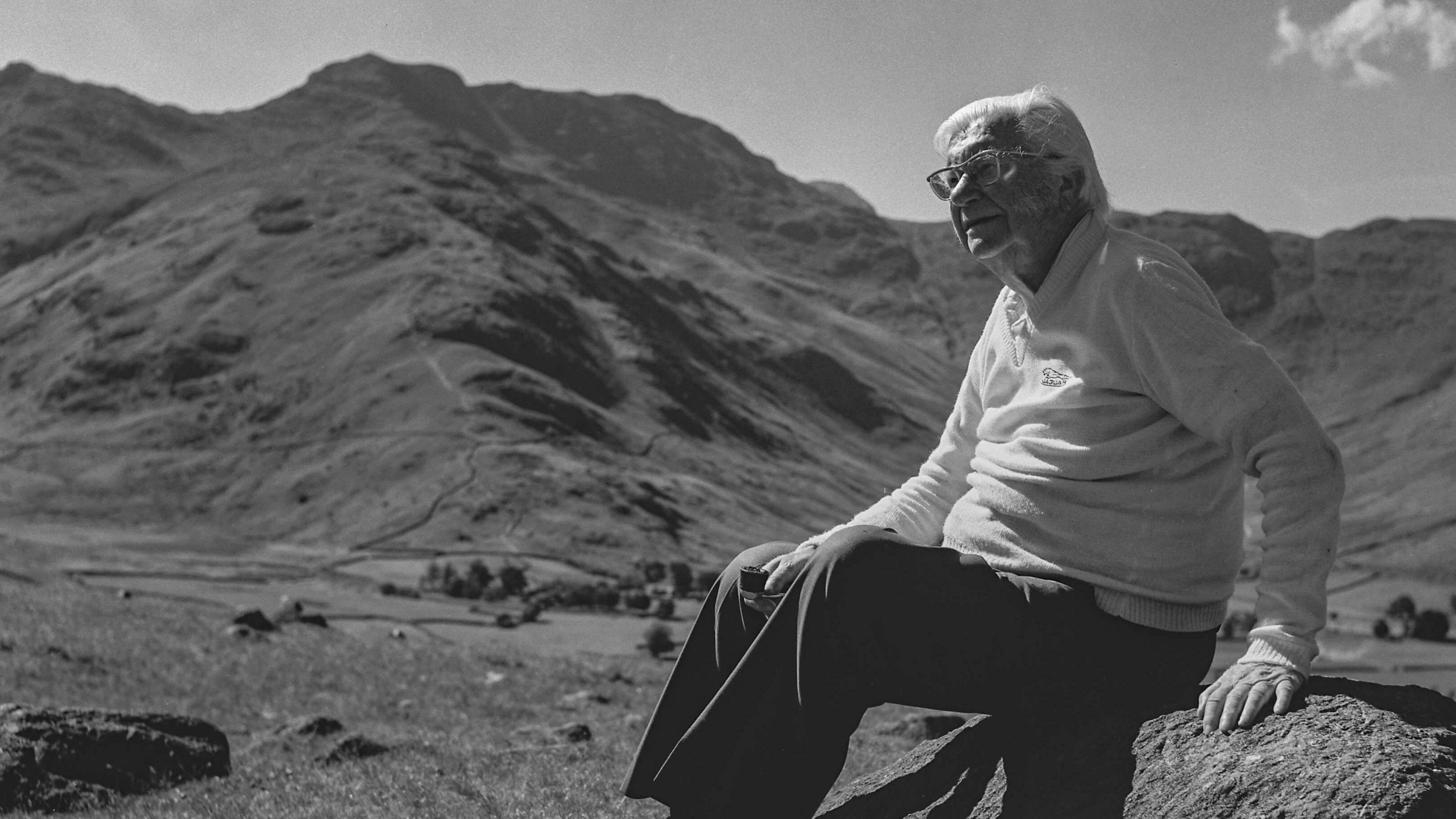 Black-and-white photograph of Alfred Wainwright sitting on a rock with the fells behind him. He is looking to the left of the image, and is wearing a light jumper and dark trousers. He has white hair and has glasses.