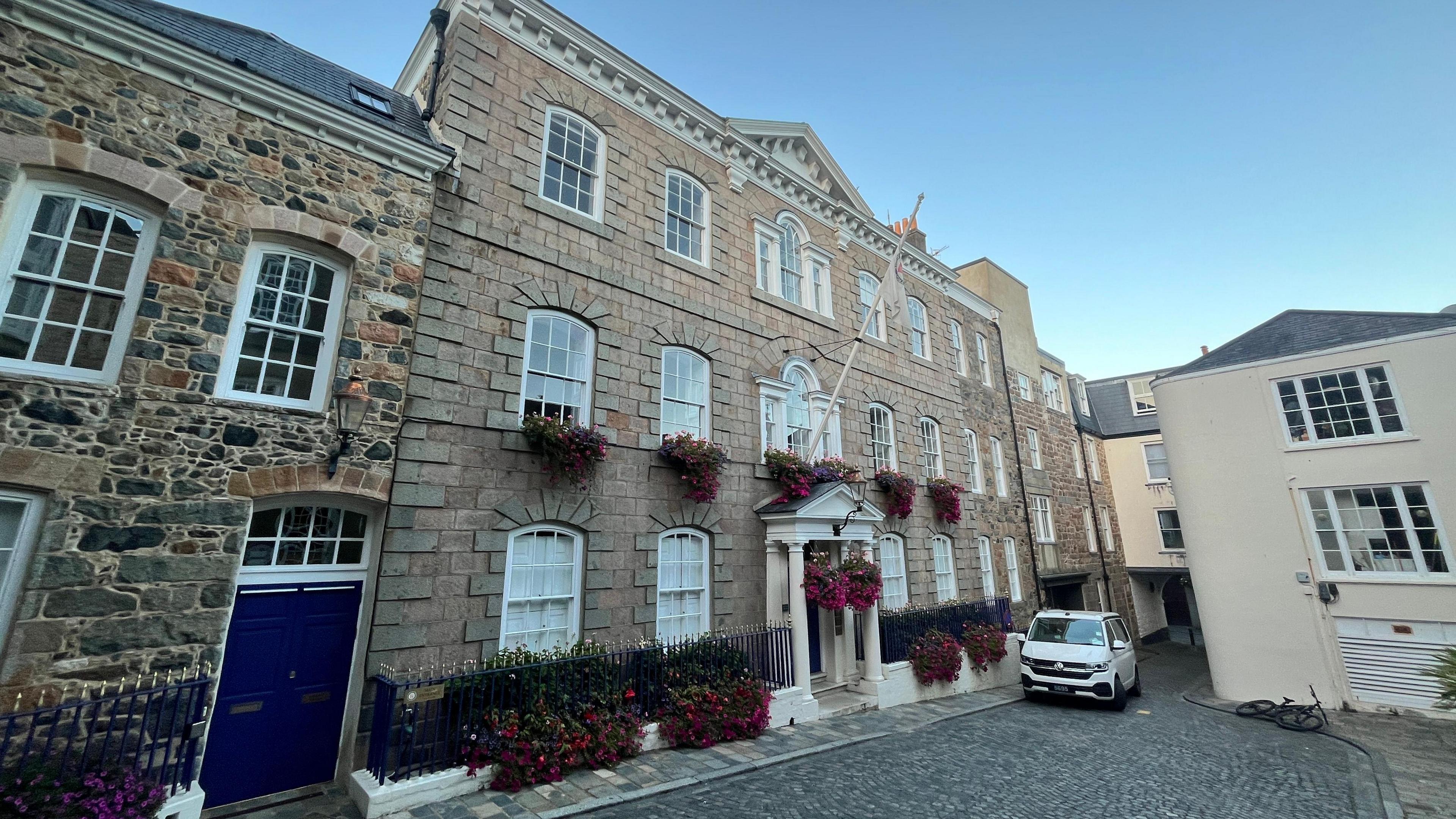 A three storey granite government building, with rows of arched windows. Handing baskets of flowers sit beneath many of them.