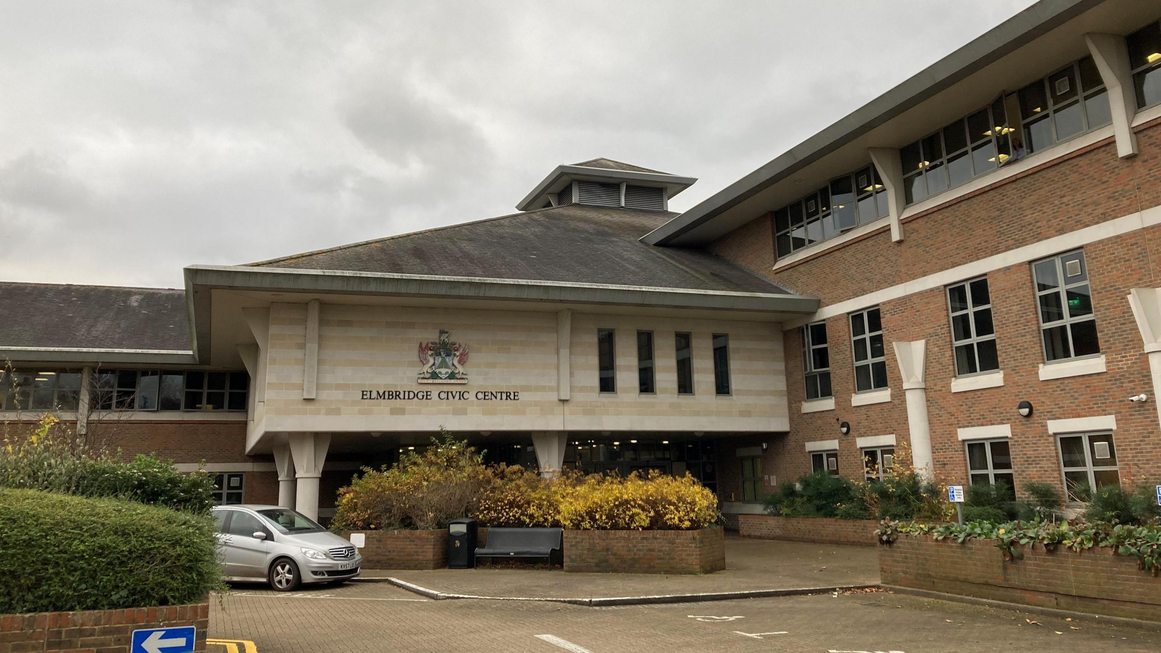 A beige and brown brick building. The building has a crest over its entrance and words reading: "ELMBRIDGE CIVIC CENTRE"