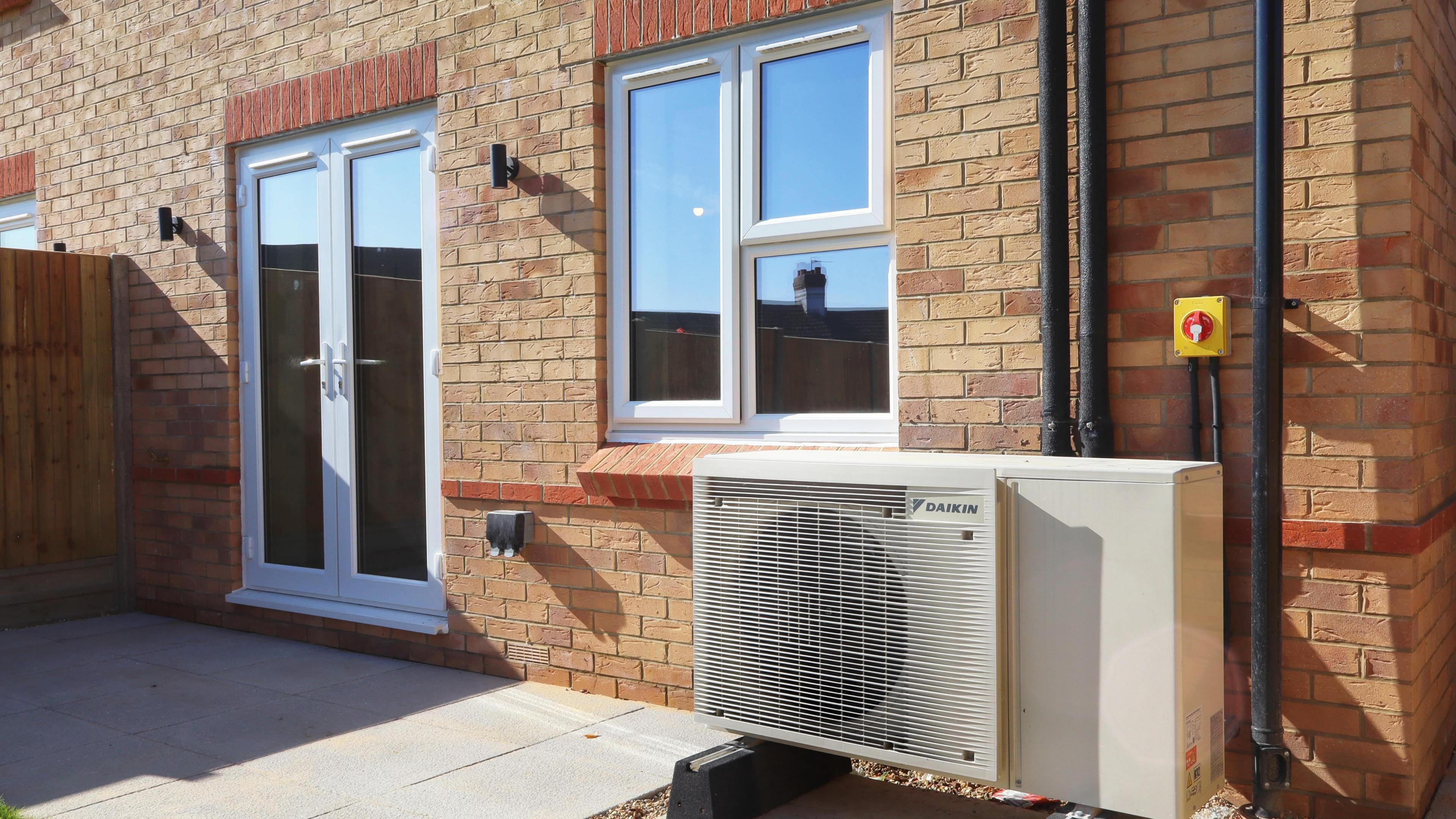 Outside of a new-build home with an air source heat pump in a large white unit with a fan grill at one side