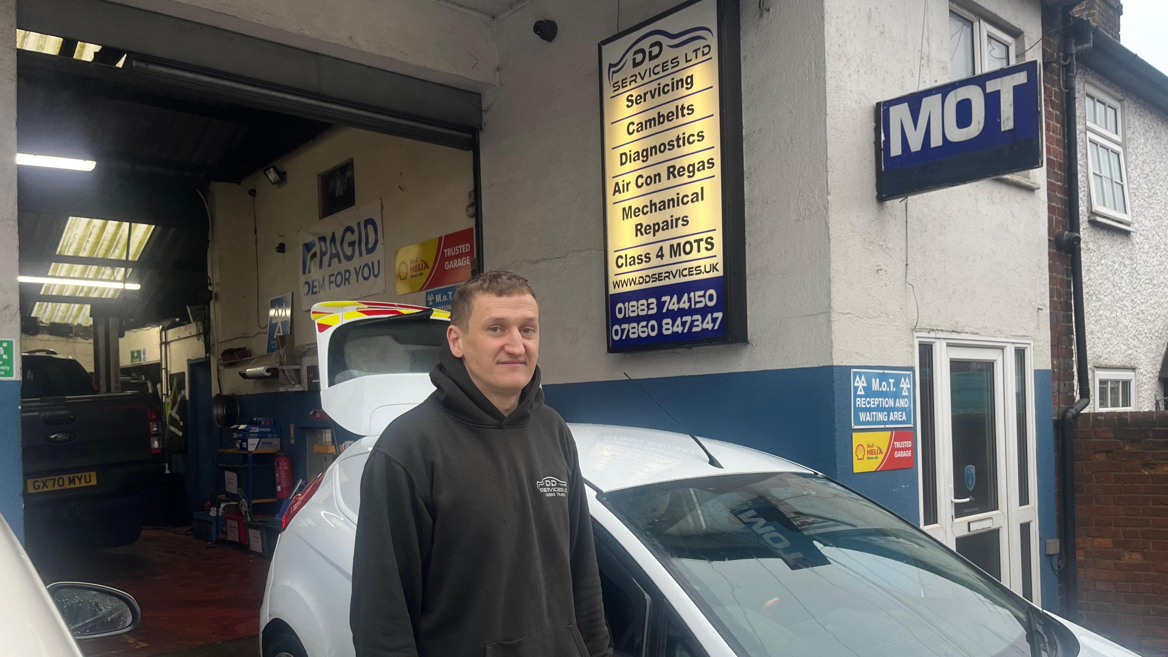 Shane Fry stands in front of his garage in Godstone, beside a white minivan, wearing a branded hoody.