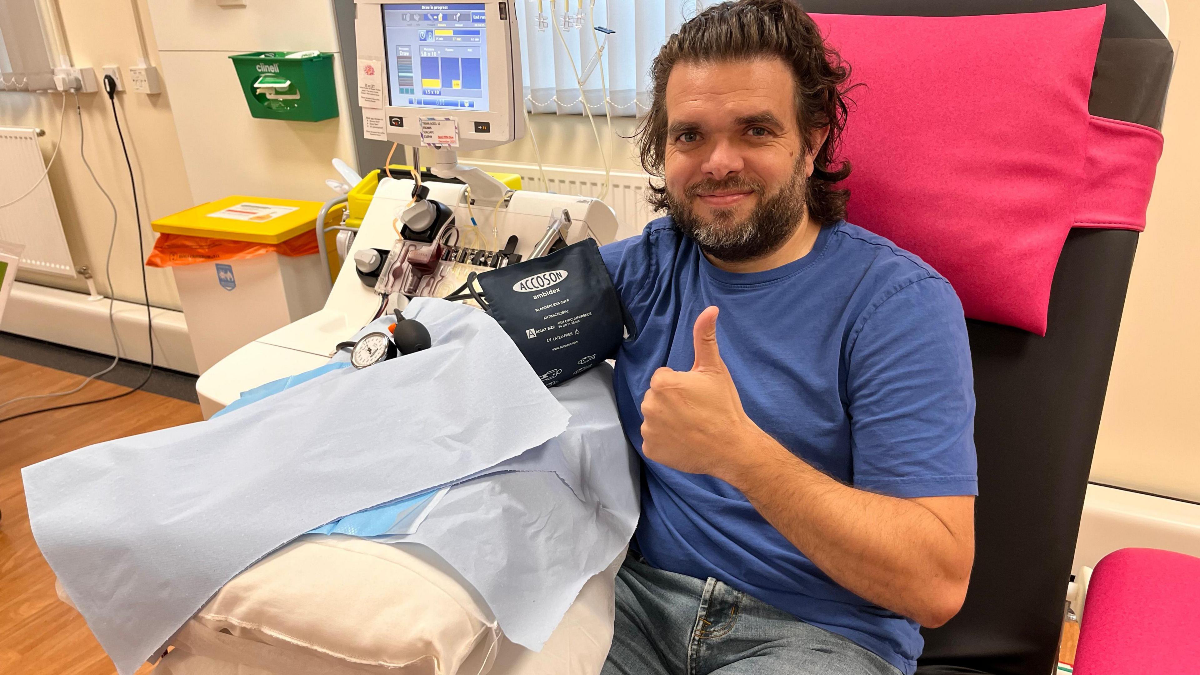 Adam mewn crys-t glas yn eistedd ar gadair ysbyty yn rhoi gwaed, gyda'i fys bawd I fyny