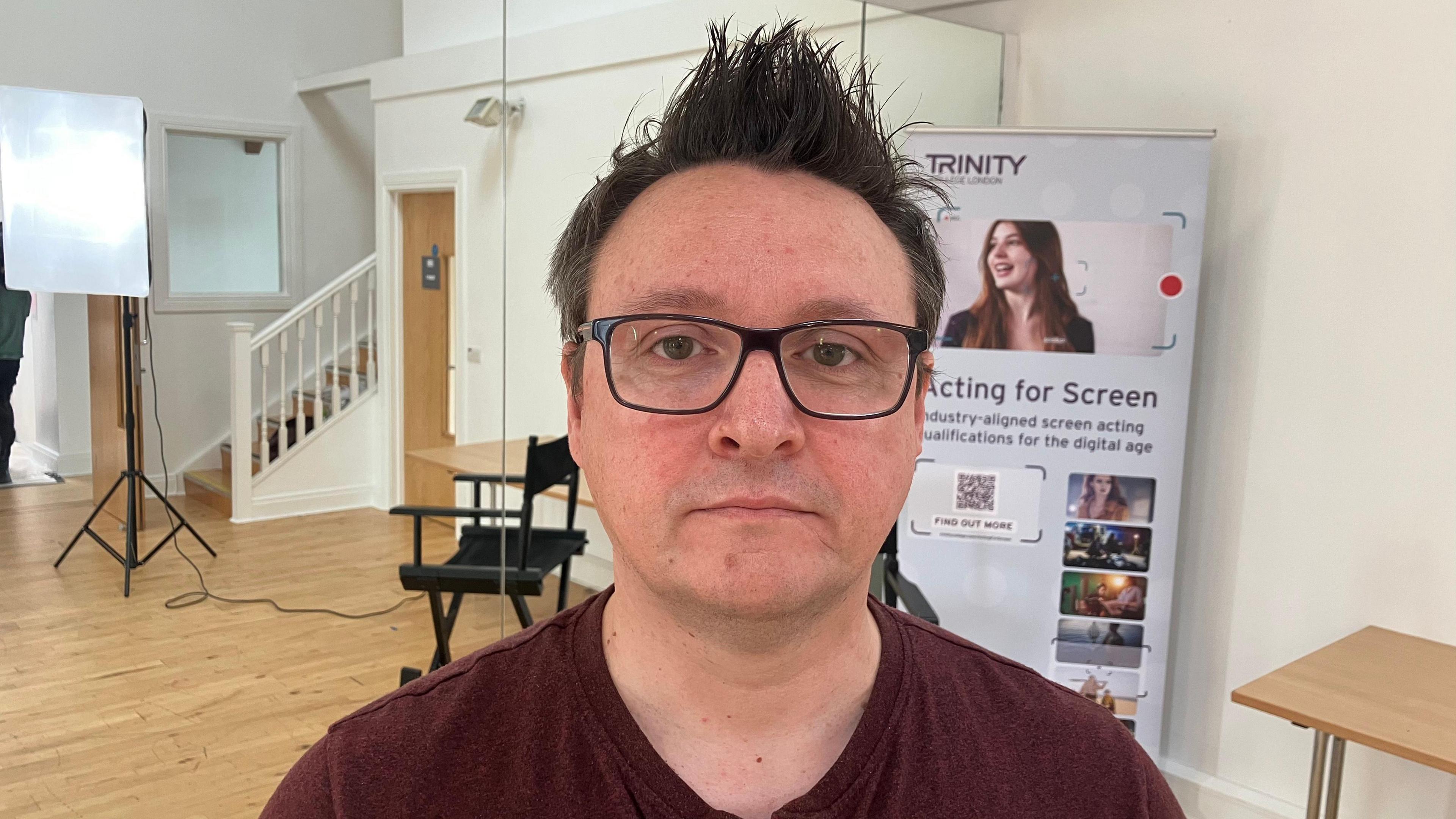 A man with spiky brown hair, wearing a burgundy t-shirt and glasses. 