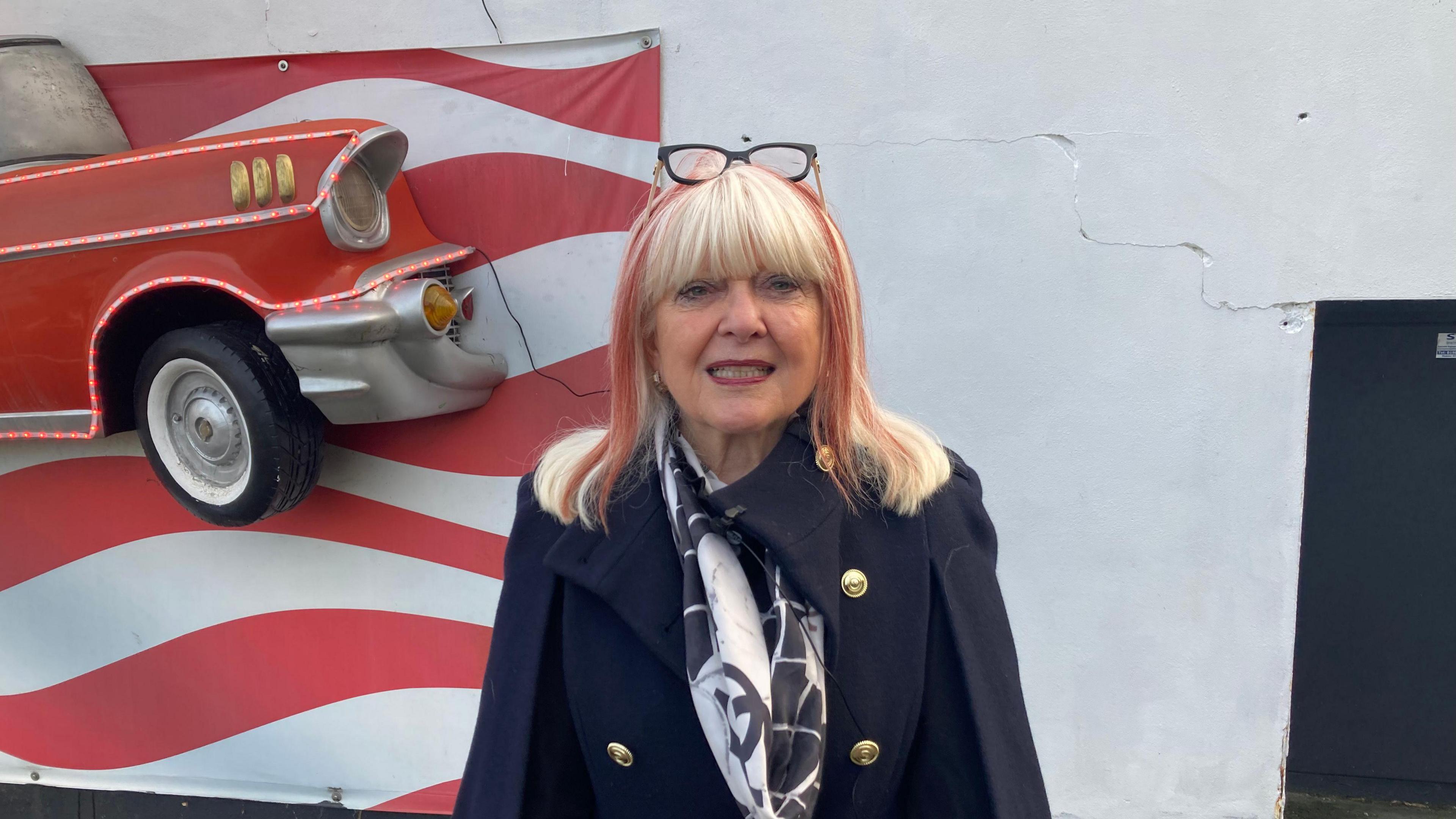 Andrea is in the centre of the frame. She has blonde shoulder-length hair, with streaks of red in it. Her glasses are on her head. She is wearing a blue coat with gold buttons. Behind her is a white wall, save for a painting of the USA flag and a classic American car embedded in the wall.