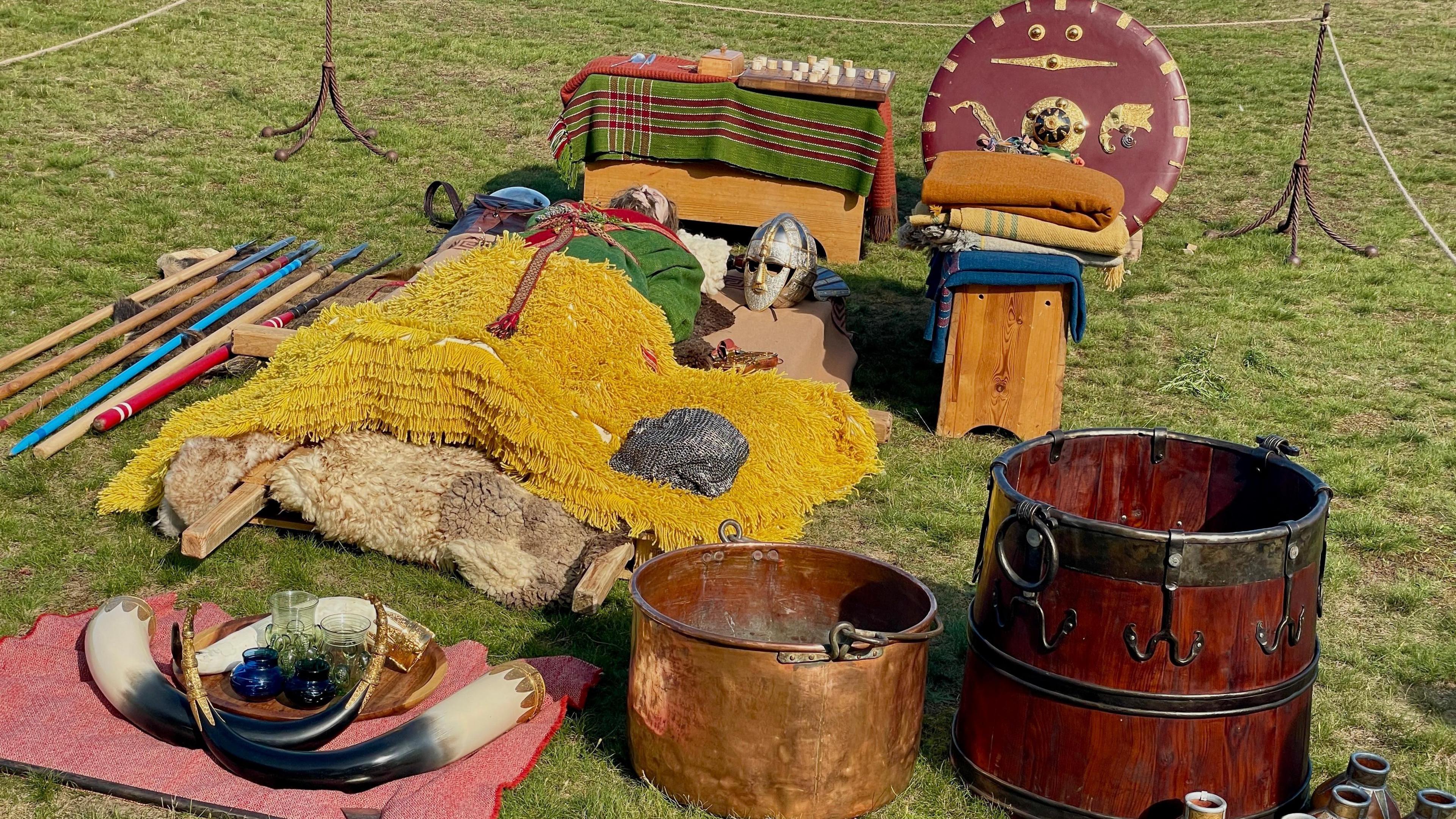 Various Anglo-Saxon items like a helmet, shield, horns and more rest on the ground.