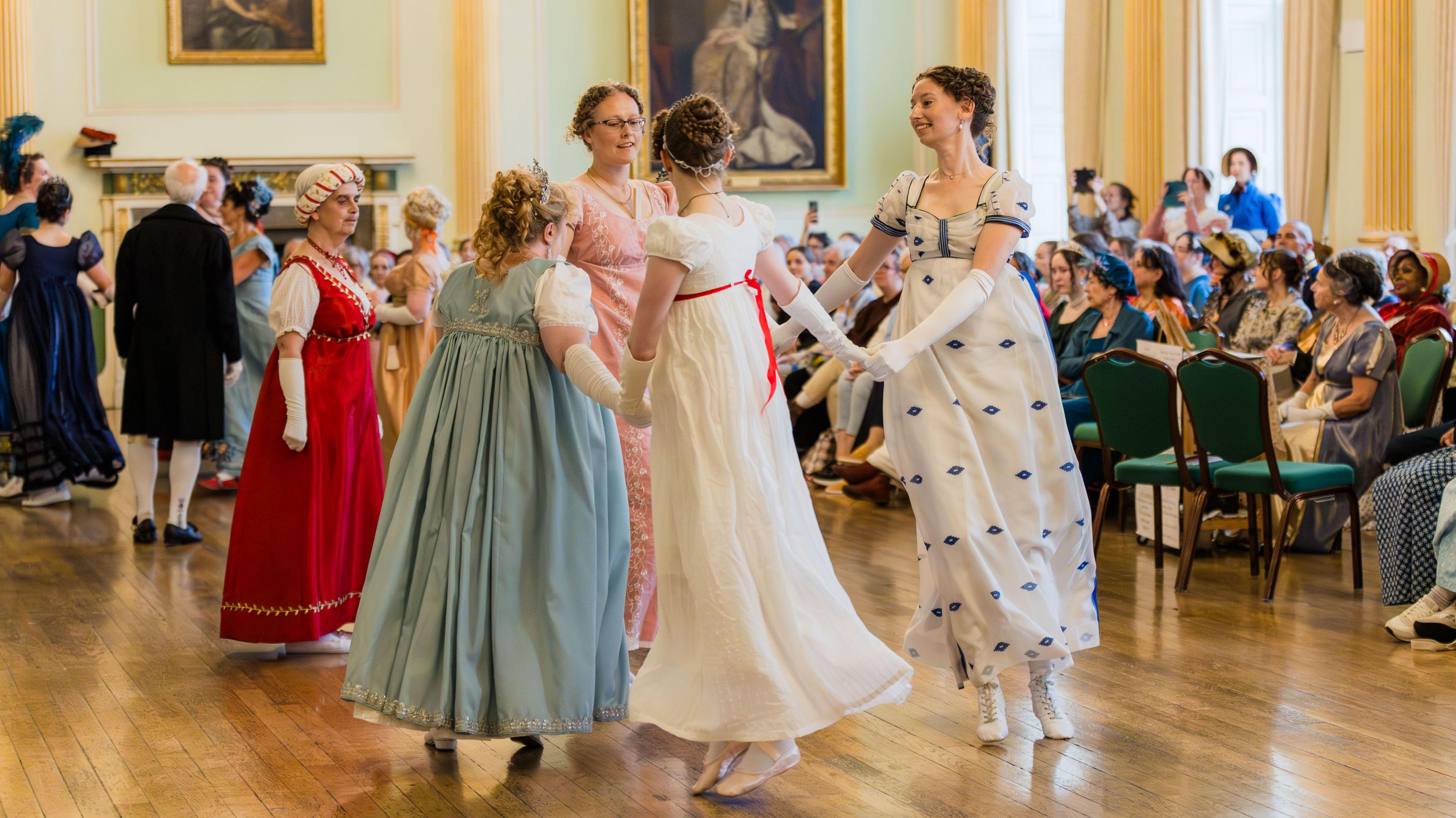 Four women dressed in regency costume dancing. They are holding hands and standing in a circle. They are in a ballroom full of others taking part in the event. 