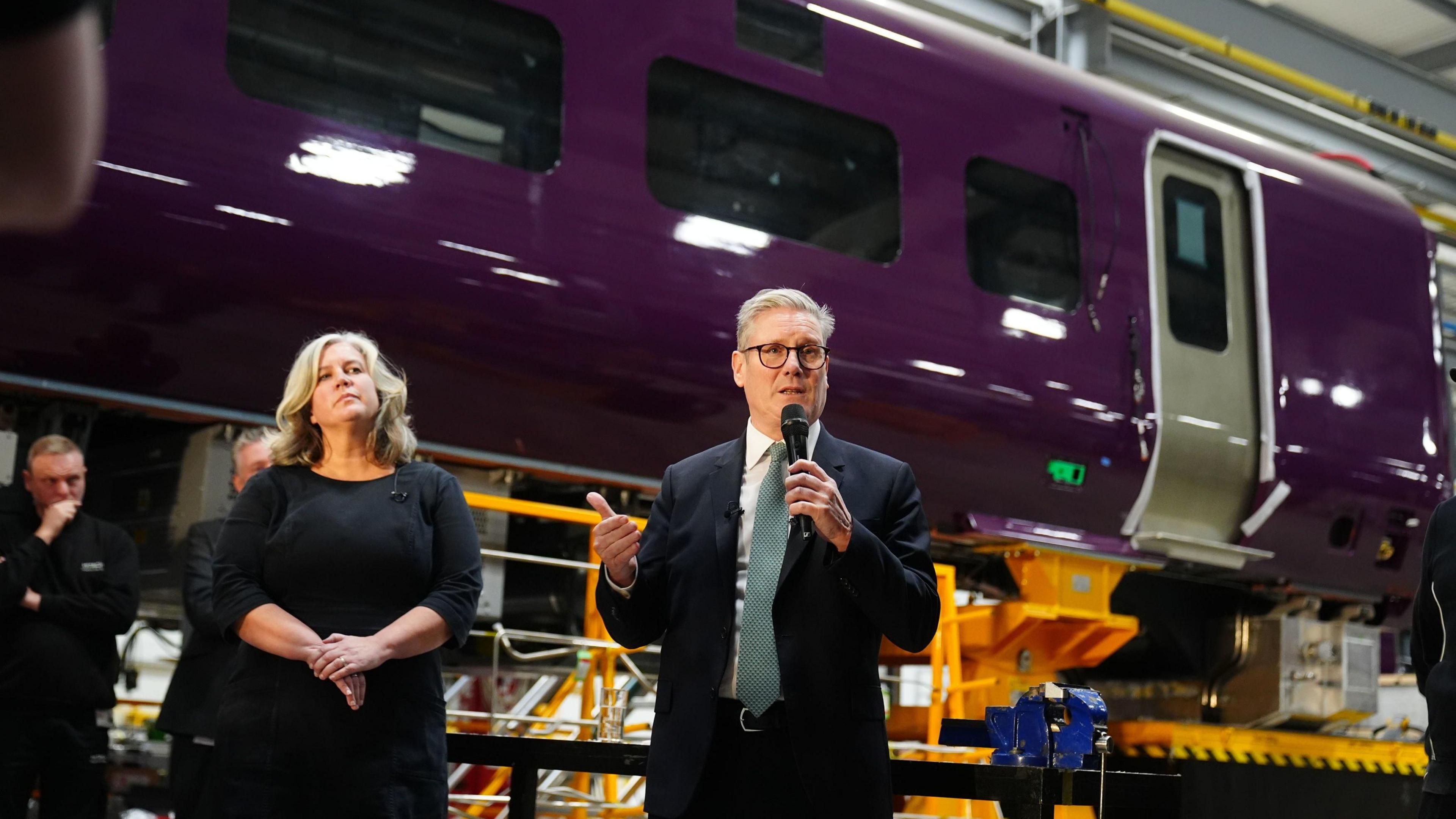 Transport Secretary Heidi Alexander (left) and Prime Minister Sir Keir Starmer, who is speaking into a microphone. A shell of train carriage is behind him, painted purple.