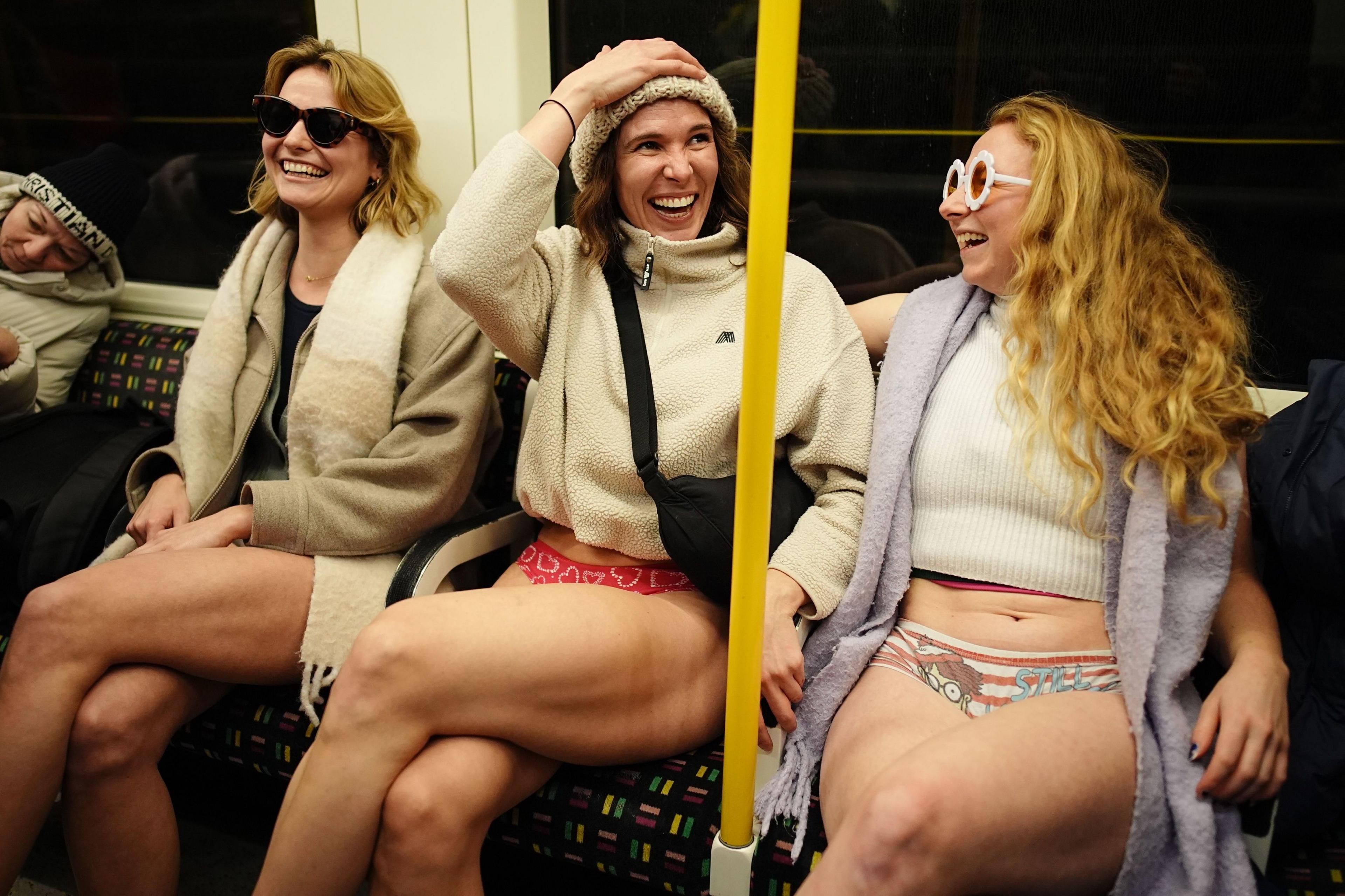 Three women laughing whilst wearing coats and jumpers but no trousers on the Tube