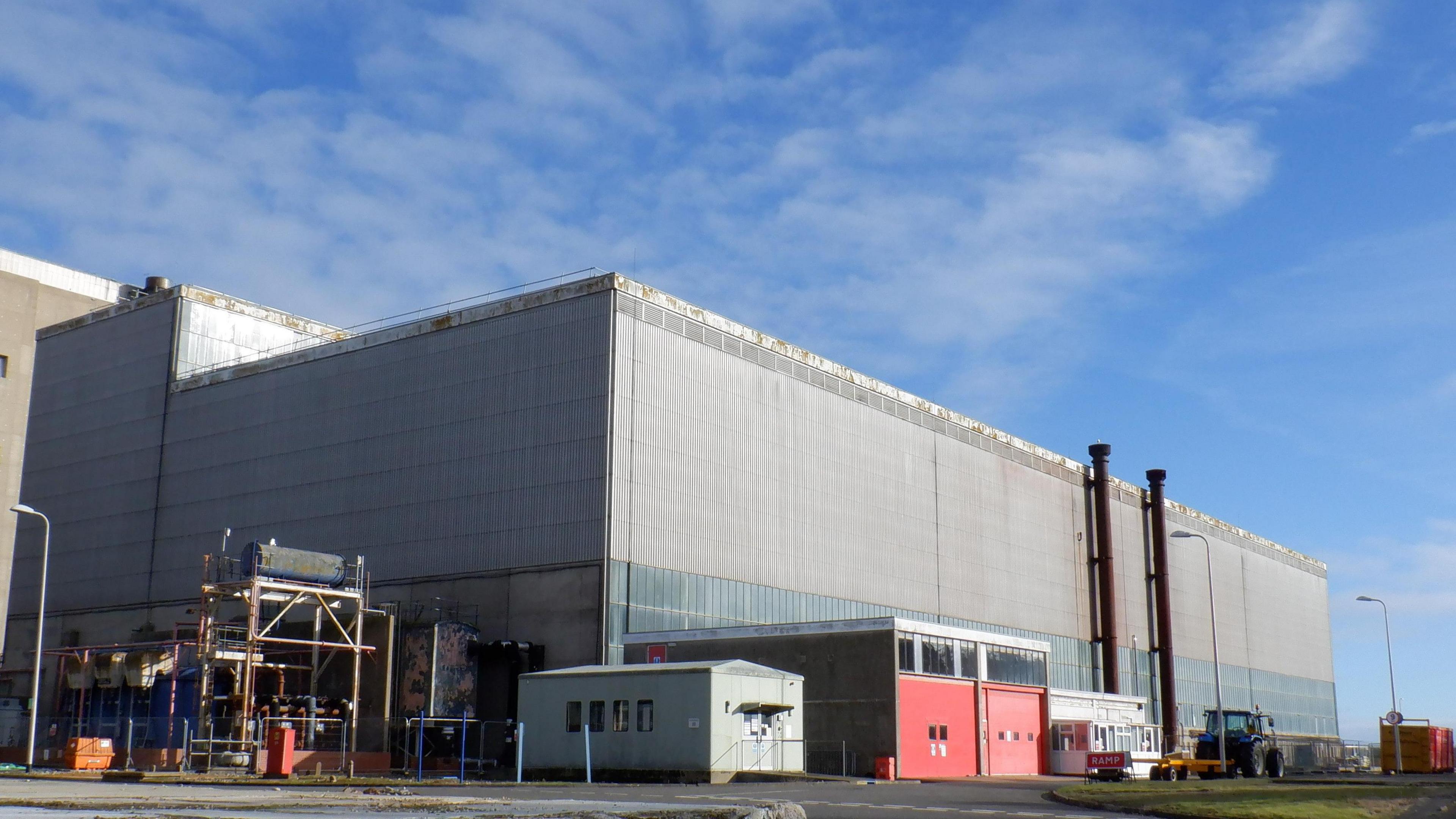 The turbine hall at Sizewell A prior to demolition works. It is a large grey building with several other small buildings surrounding it. It has been pictured on a sunny, blue day.