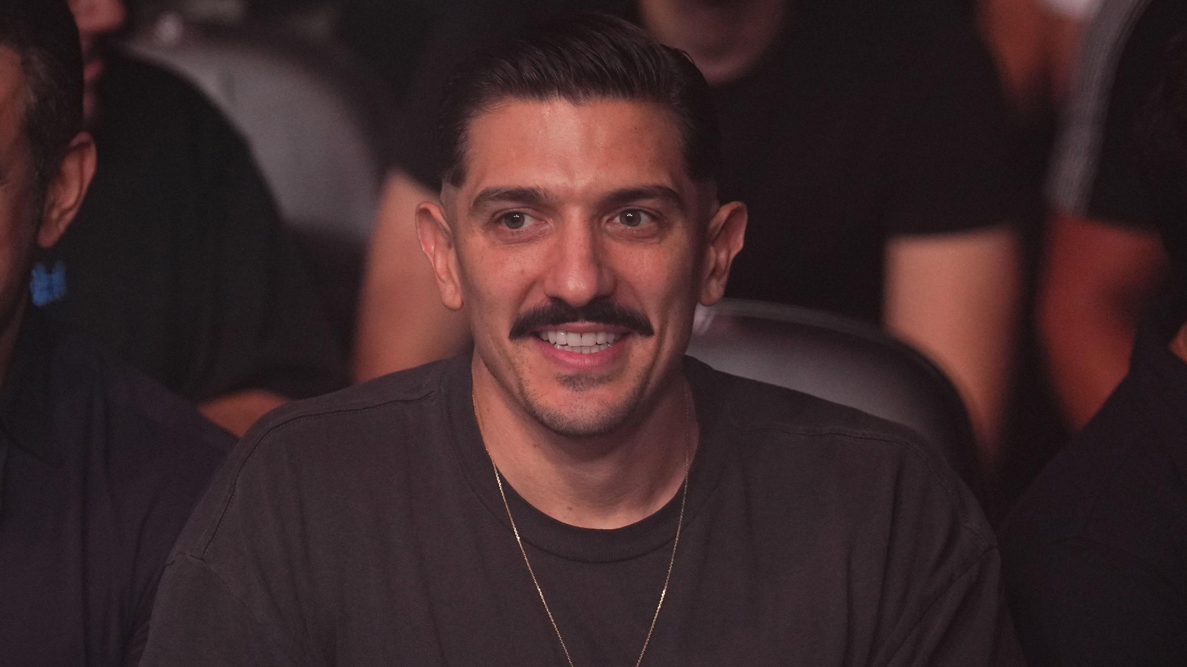 Andrew Schulz, pictured in 2021 at a UFC event. Andrew has short dark hair, brown eyes and a brown moustache. He wears a gold chain over a dark grey T-shirt and is pictured sitting at the event, with audience members blurred behind him. 