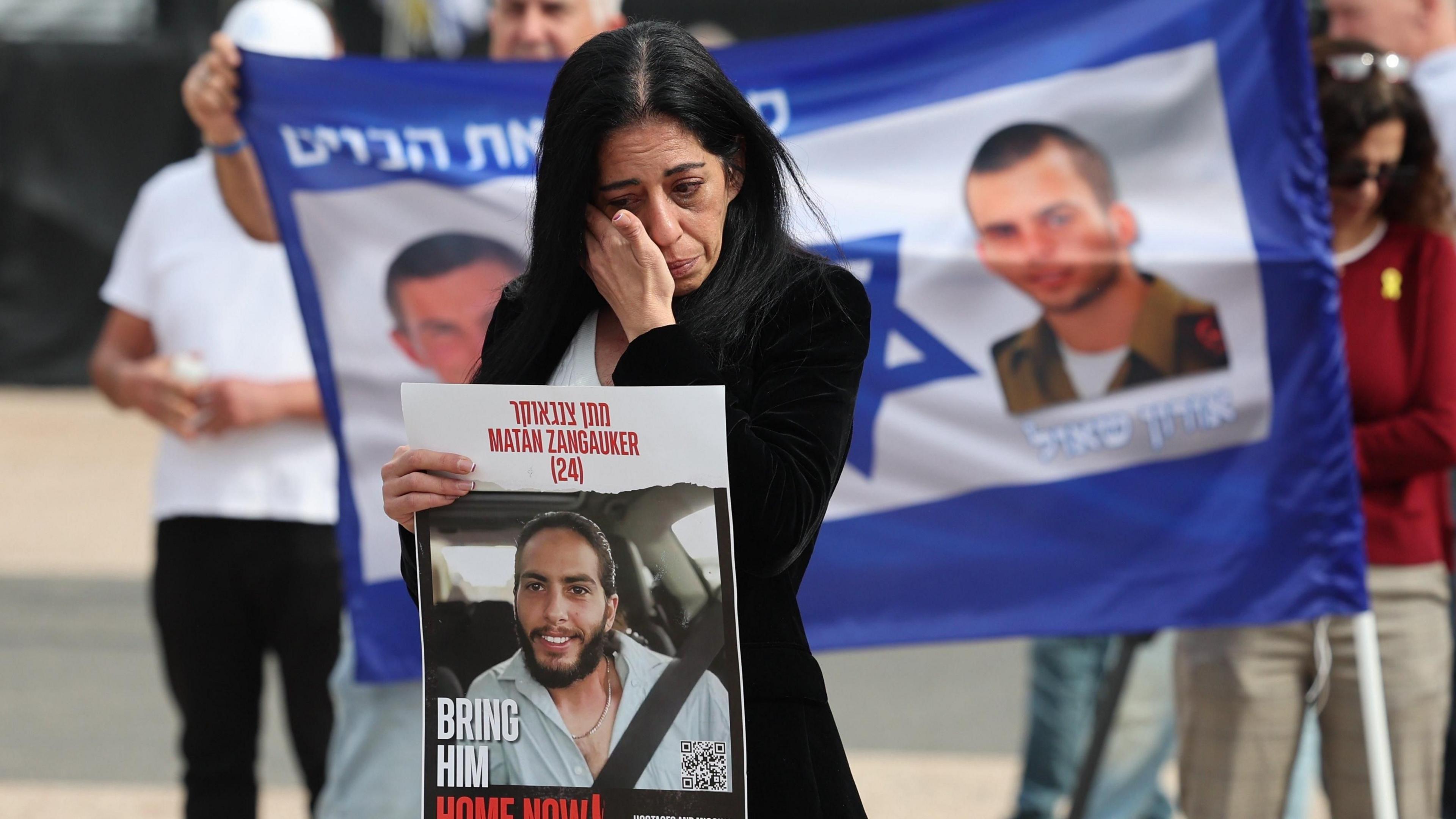 Einav Zangauker, the mother of Israeli hostage Matan Zangauker, holds a poster of her son reading "Bring him home now!", during a press conference outside the Israeli military's headquarters in Tel Aviv, Israel, while behind her people hold a banner with a Star of David and two photos