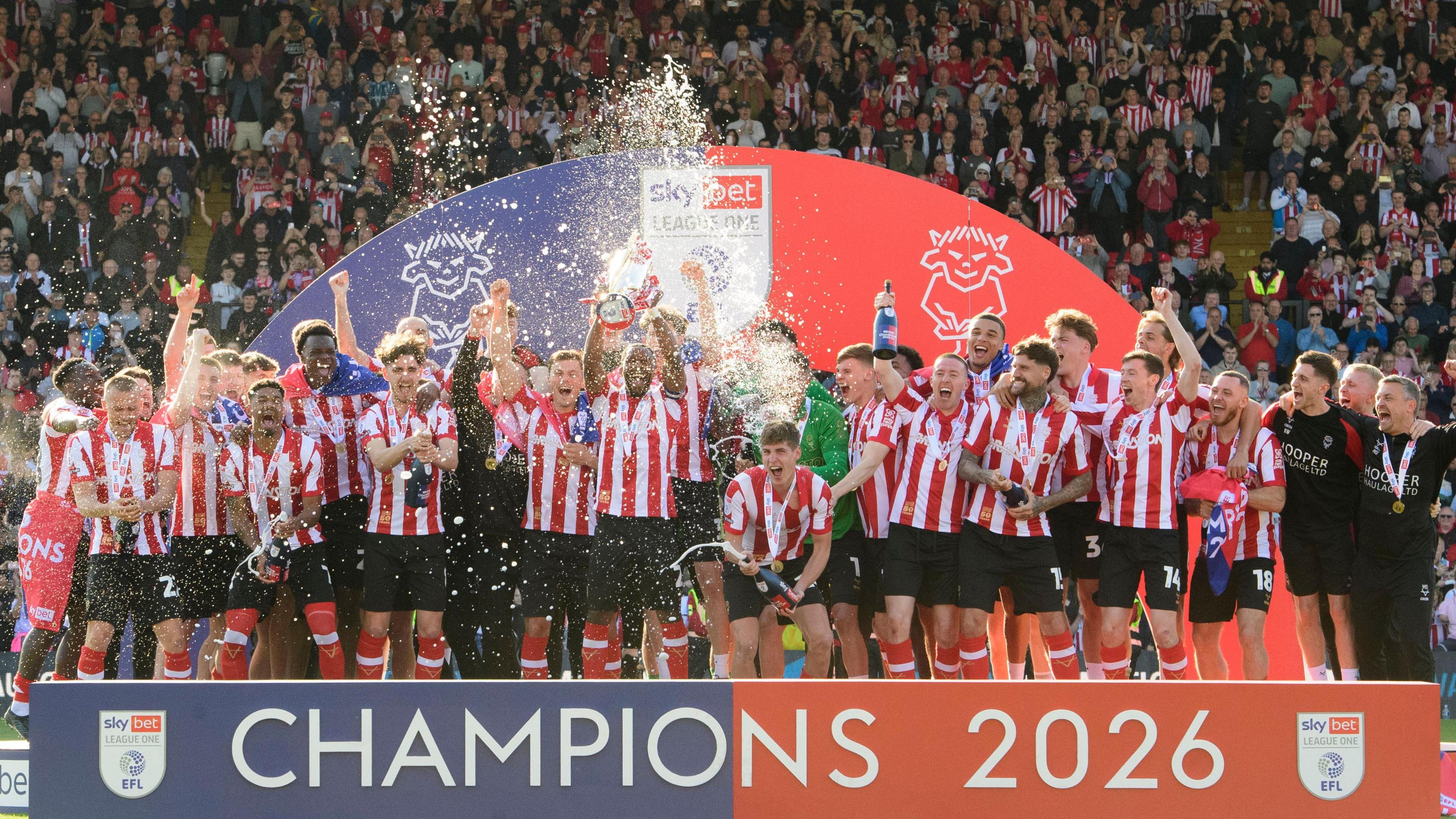 Lincoln City's players, wearing their red and white striped home shirt with black shorts and red socks, lift the League One trophy. Champagne can be seen getting sprayed into the air. The fans can be seen behind the stand which says "CHAMPIONS 2026" on it.
