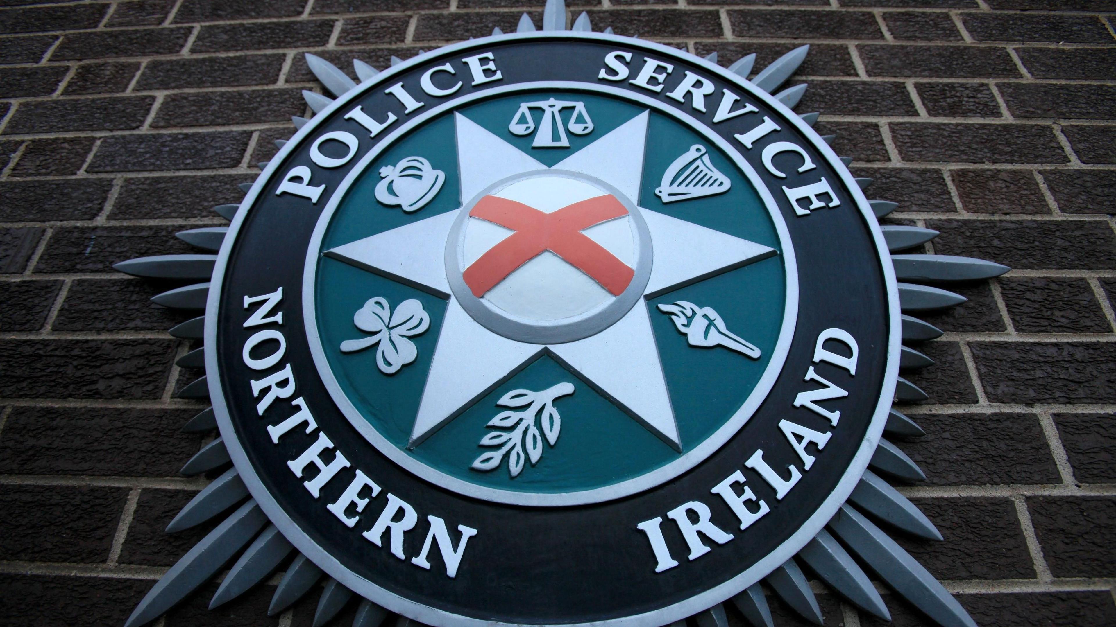 A close up of the PSNI emblem on a brick wall. It consists of a star shape in the centre, with the scales of justice, a harp, a torch, a leaf, a shamrock, and crown.