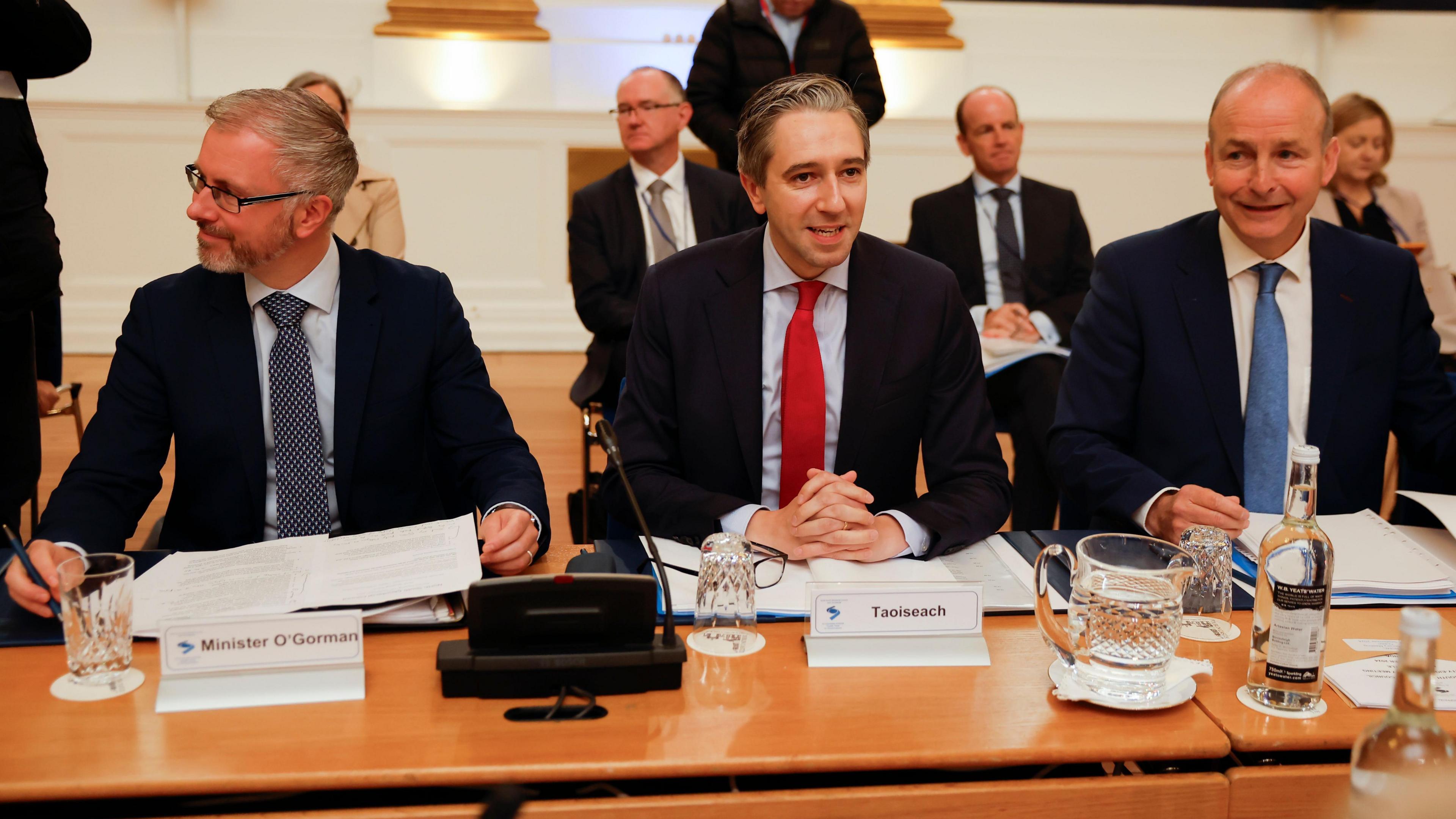 Three men - Roderic O'Gorman, Simon Harris and Micheál Martin - sit together at a large table. On the left, O'Gorman is wearing a suit and tie. He has grey hair, is wearing rectangle shaped glasses and is looking to the side. Harris sits in the middle and is mid-speech, wearing a suit and tie. Martin is sat on the right and smiles while wearing a suit and tie.