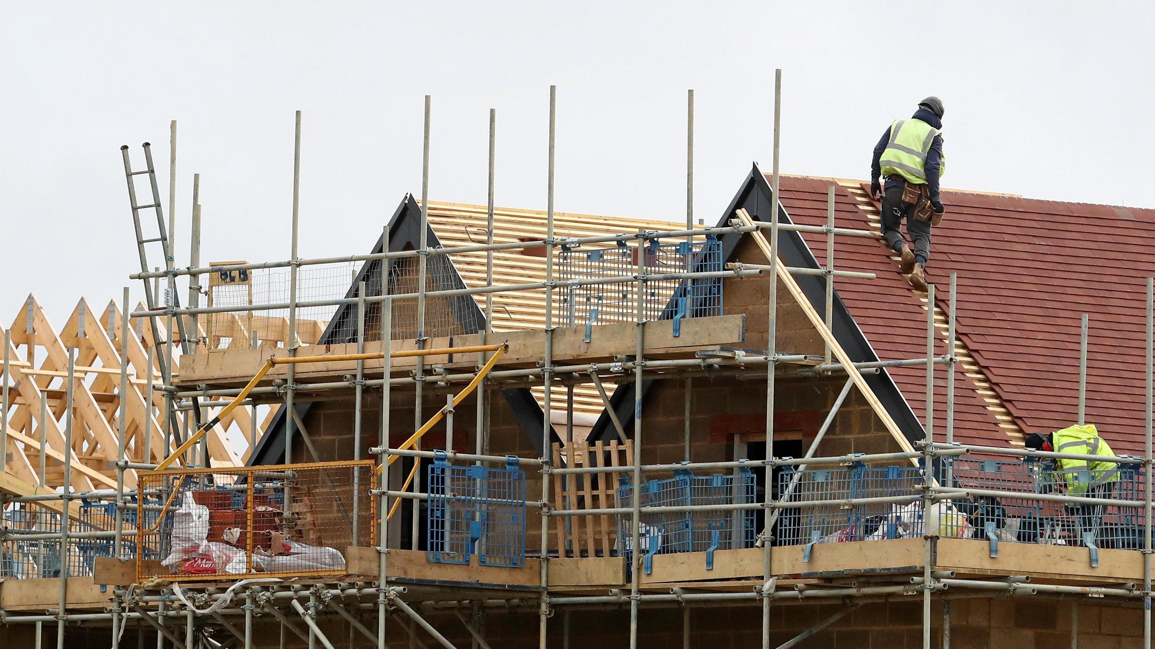 Construction workers tile a roof on a new build housing estate with unfinished homes covered in scaffolding.